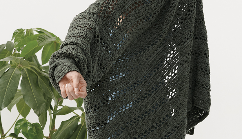 Häkelanleitung Cardigan aus Creative Sport dk: Dunkler, gehäkelter Cardigan mit offenem Muster, getragen vor einer Pflanze.