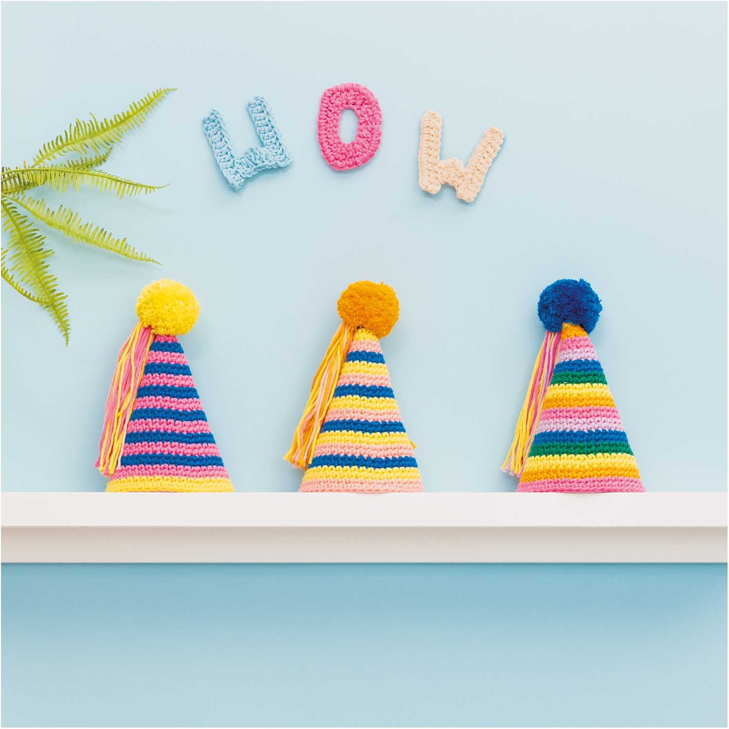 Creative Cotton aran: Drei bunte, gestrickte Partyhüte mit Pompons auf einem Regal vor hellblauer Wand.