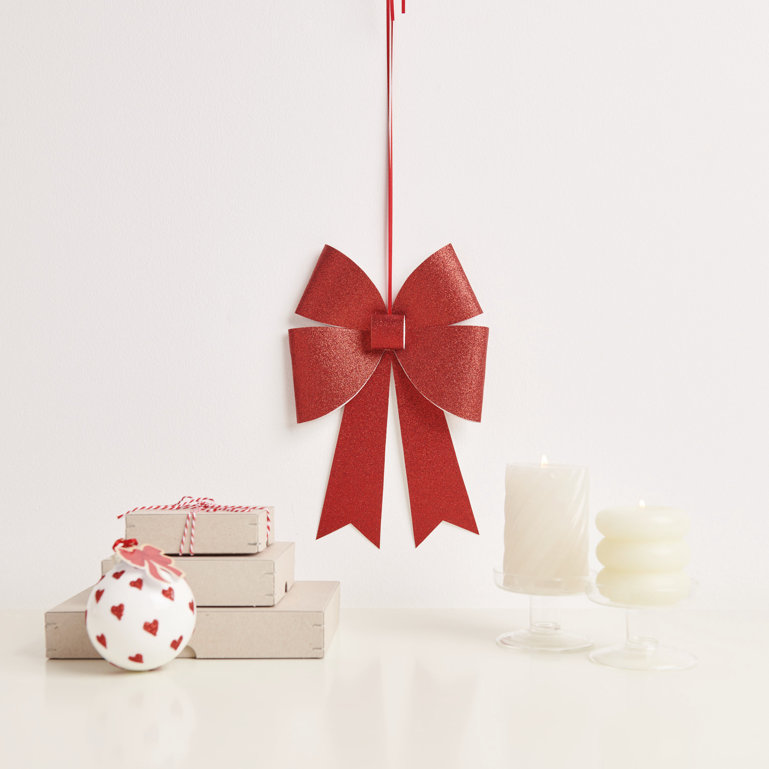 Paper Poetry Bastelset XL-Schleife Glitter: Eine dekorative rote Schleife hängt neben Kerzen und Geschenken. Perfekt für festliche Anlässe.