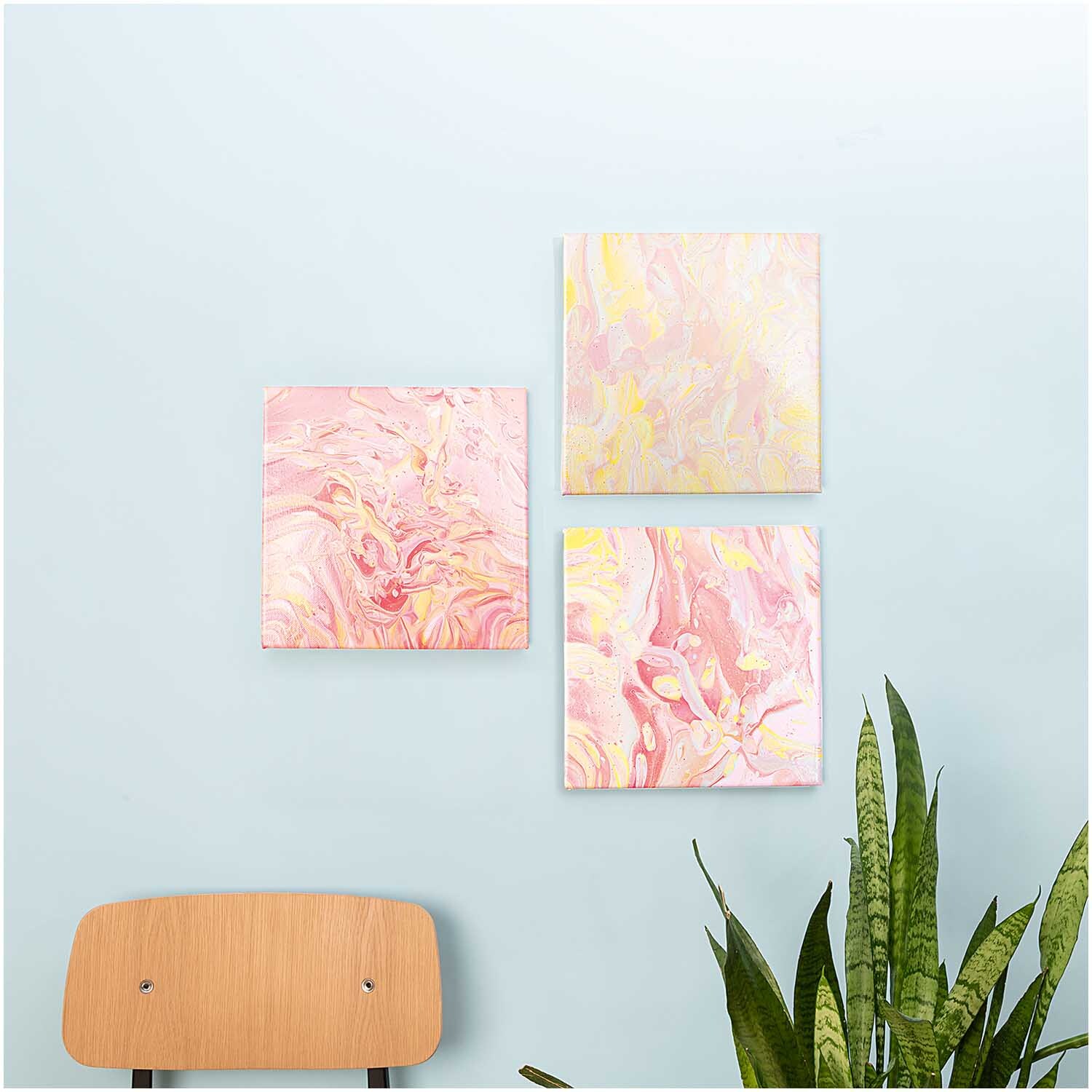 Pouring Medium seidenglänzend von Rico Design, transparent, auf drei abstrakten Leinwänden in Pastelltönen an einer hellblauen Wand.