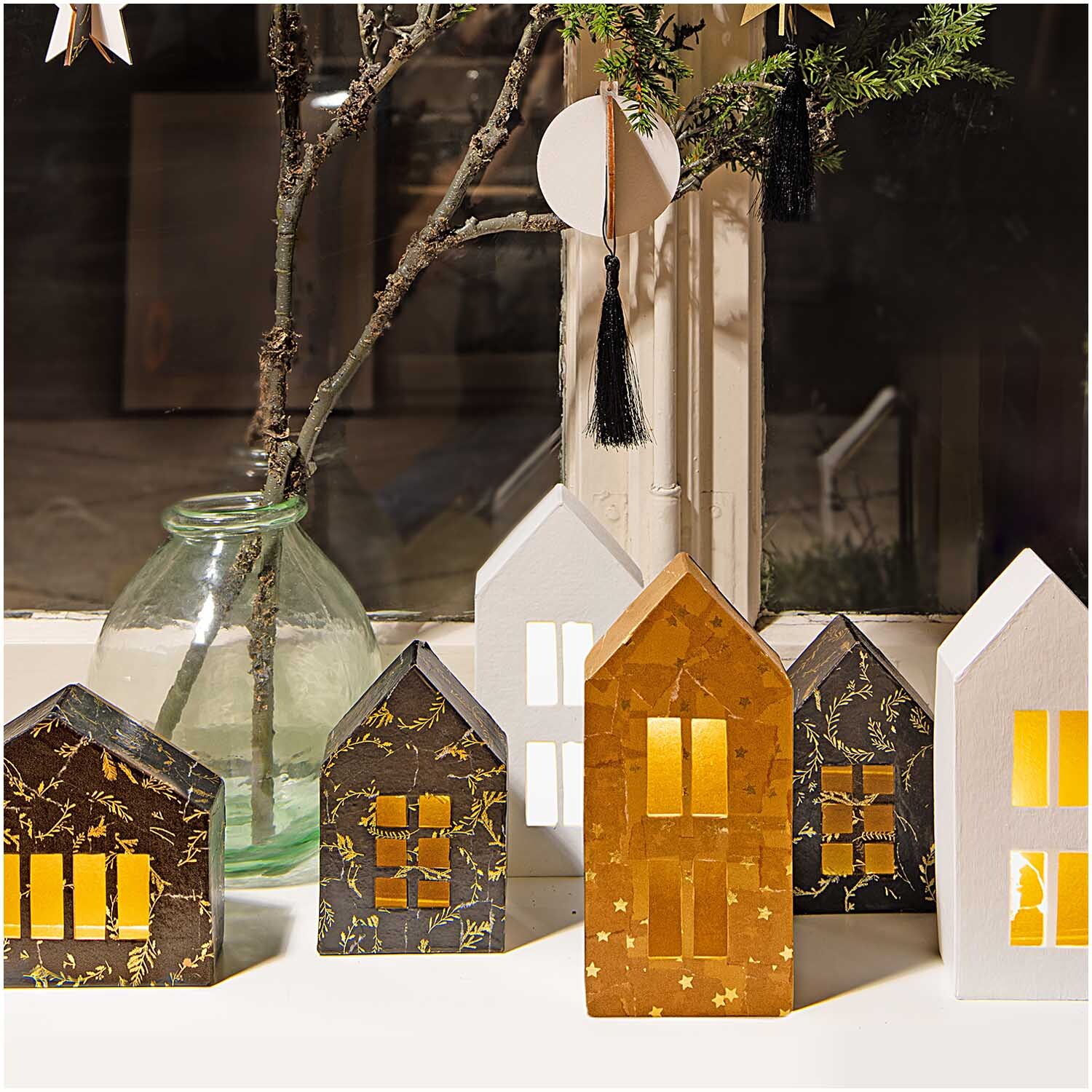 Paper Patch Papier Nostalgic Christmas Kränze zeigt dekorative Häuser mit leuchtenden Fenstern vor einem Fenster.