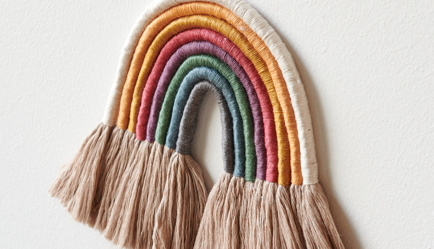 Bastelanleitung Regenbogen mit Sticktwist gestalten: Handgemachter Regenbogen aus buntem Garn an einer Wand.