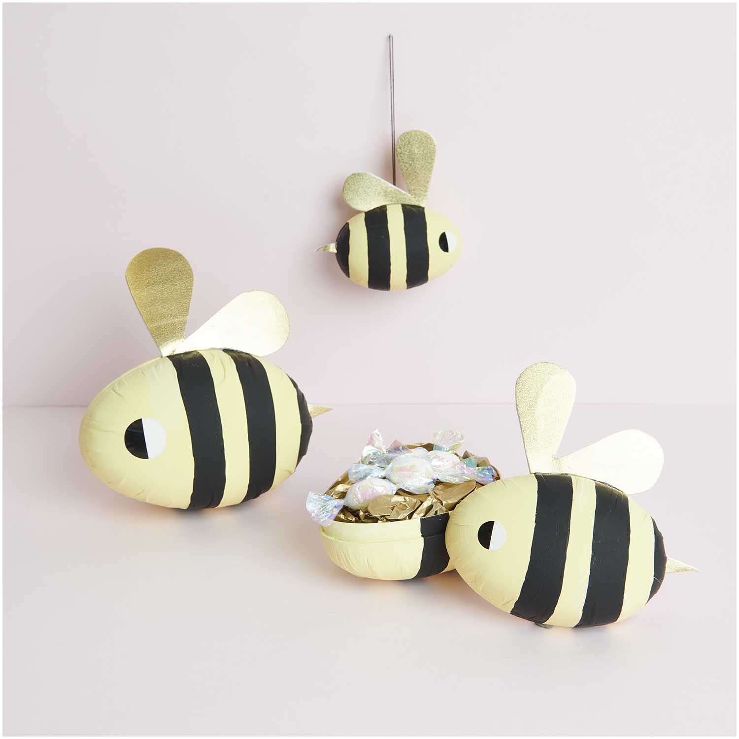 Pappmaché Eier weiß 9cm, 12cm & 15cm von Rico Design. Drei bienenförmige Dekorationen in Schwarz-Gelb auf rosa Hintergrund.