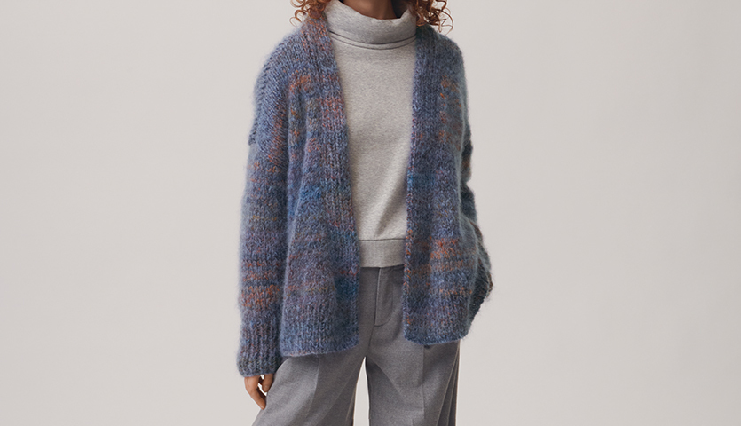 Strickanleitung Zopf-Jacke aus Creative Mohair Melange: Blaue, gemusterte Jacke über grauem Rollkragenpullover, kombiniert mit grauer Hose.