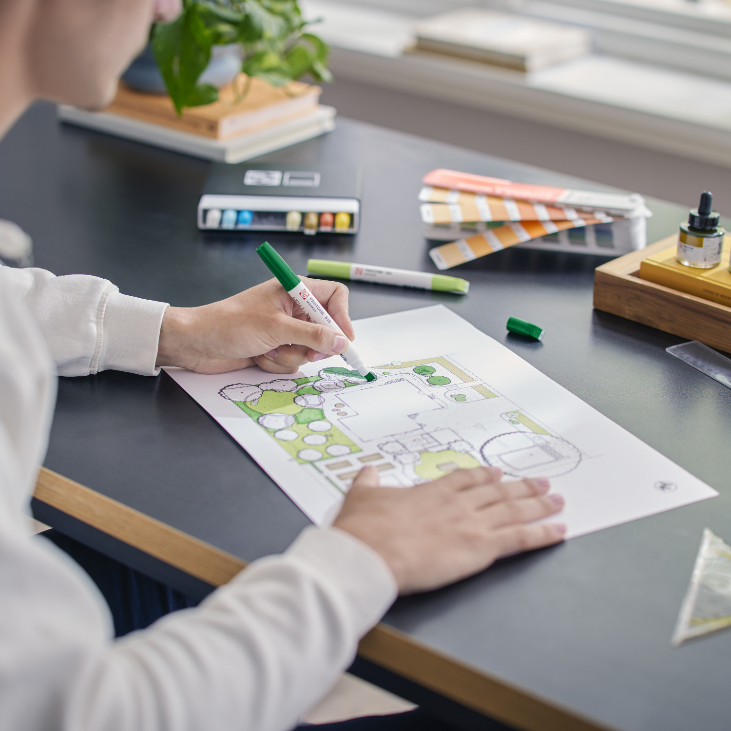 Talens | Pantone Marker Set natur, 9-teilig, zeigt eine Person beim Zeichnen eines Landschaftsdesigns mit grünen Markern auf einem Tisch.
