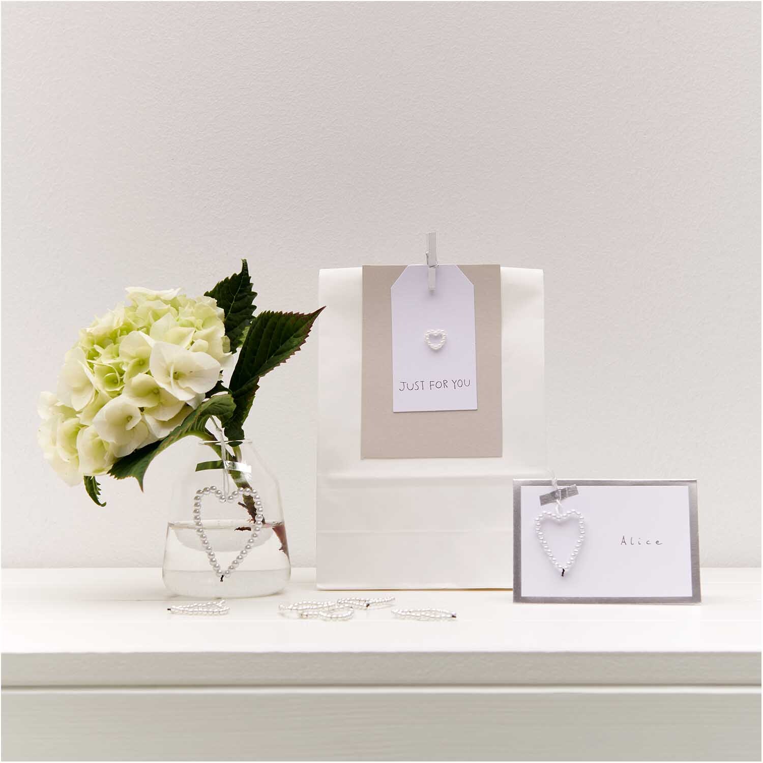 Perlenhänger Herz klein perlmutt von Rico Design: Eleganter Herzanhänger neben Blumen und Geschenktüte auf weißem Tisch.
