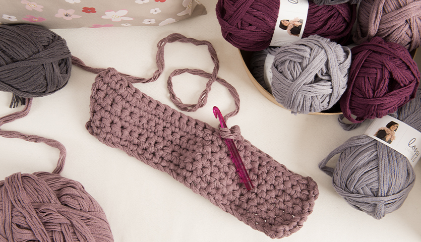 Häkelanleitung Creative Loopy Platzsets: Rosa Häkelstück mit Häkelnadel, umgeben von Garn in verschiedenen Lilatönen.