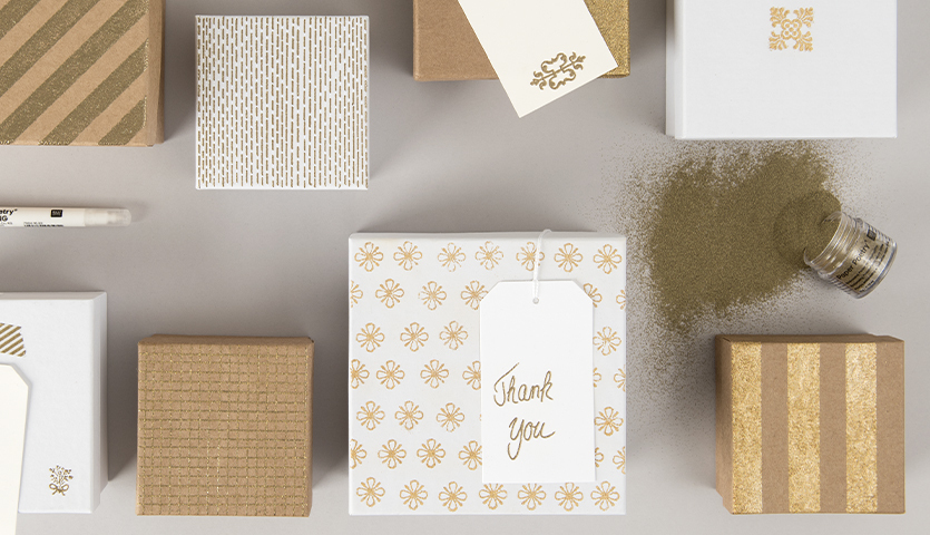 Anleitung Embossing: Verschiedene Geschenkboxen mit goldenen Mustern und Embossing-Pulver auf grauem Hintergrund.