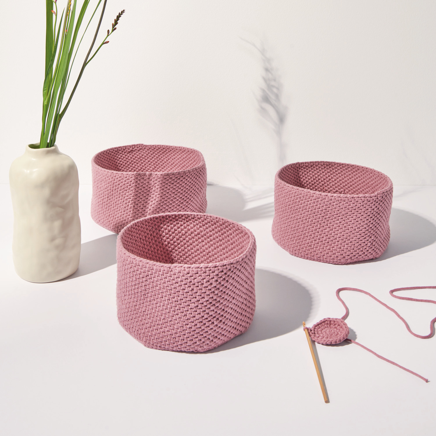 Häkelset Korb Modell 04 aus Boho Crochet: Drei rosa Körbe neben einer Vase, mit Häkelnadel und Garn.
