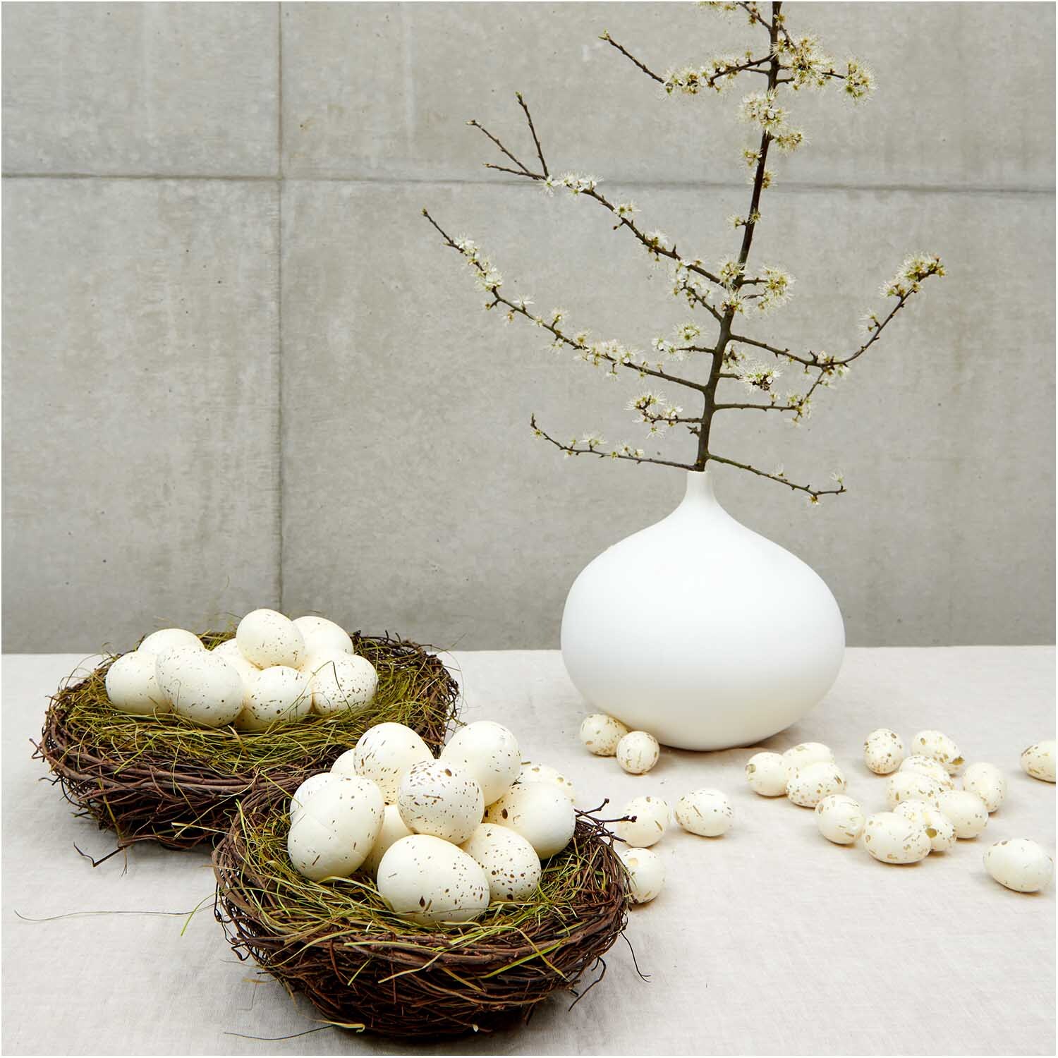 Naturnest mit Ostergras 20cm, dekoriert mit weißen Eiern, neben einer Vase mit blühendem Zweig.