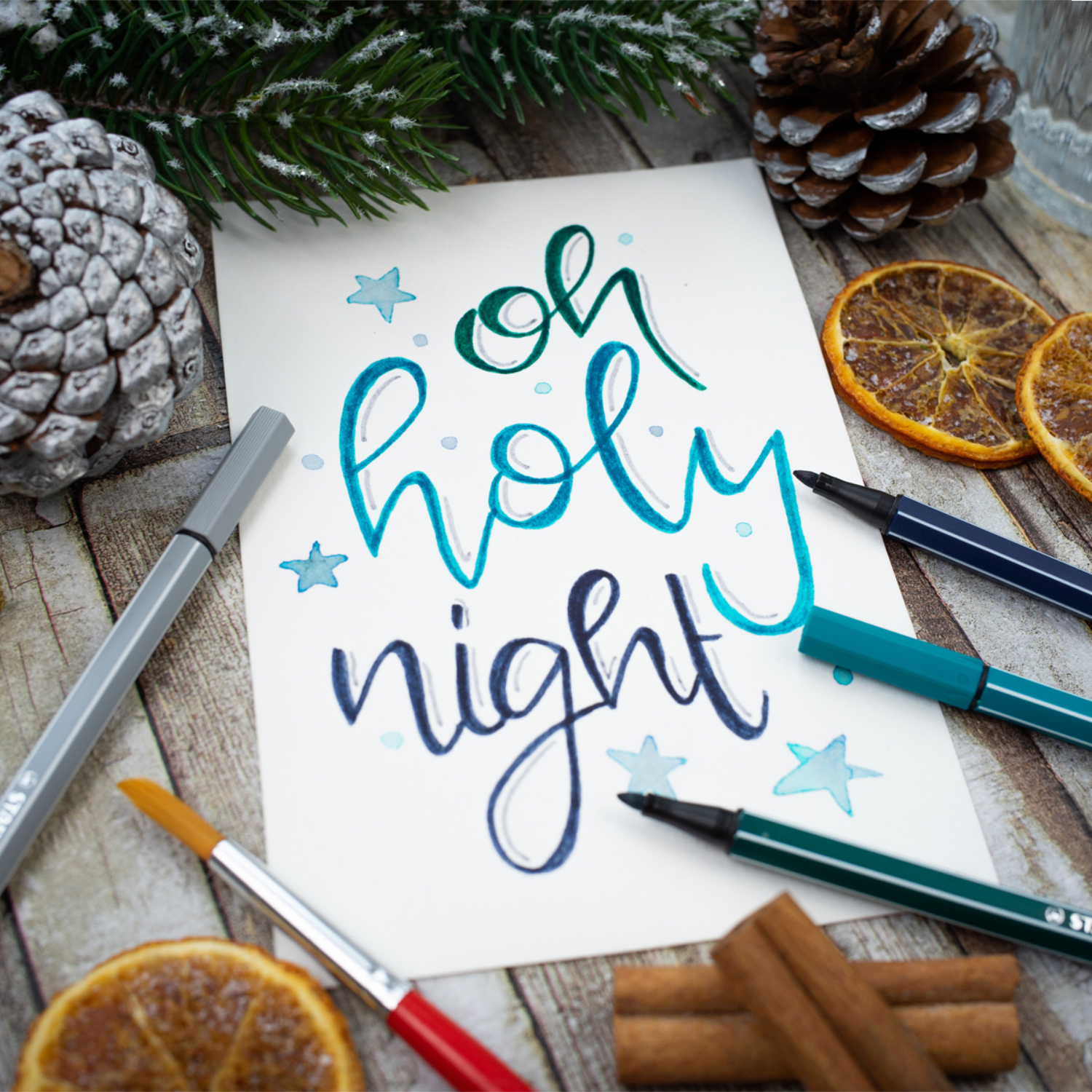 Pen 68: Handgeschriebener Text "oh holy night" mit STABILO Stiften, umgeben von Tannenzapfen und getrockneten Orangen.