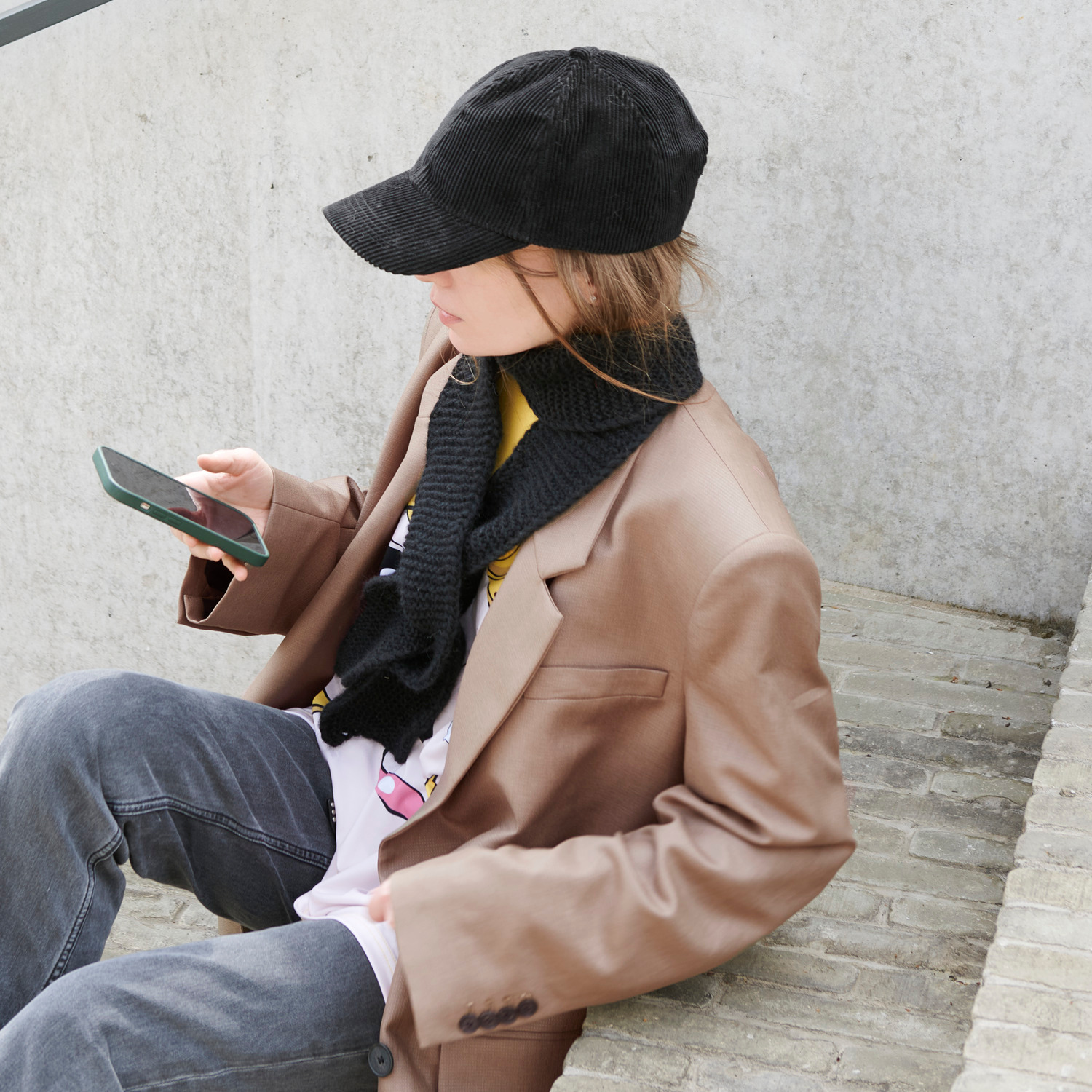 Strickset Mini Schal Modell 03a: Person in braunem Blazer und schwarzem Schal, sitzt und schaut auf ein Smartphone.