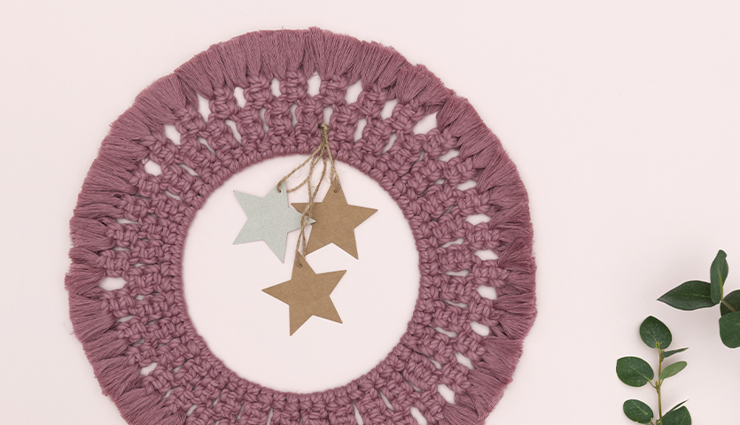 Anleitung Makramee-Weihnachtsdeko: Ein rosafarbener Makramee-Kranz mit Sternanhängern, dekoriert mit grünen Blättern.