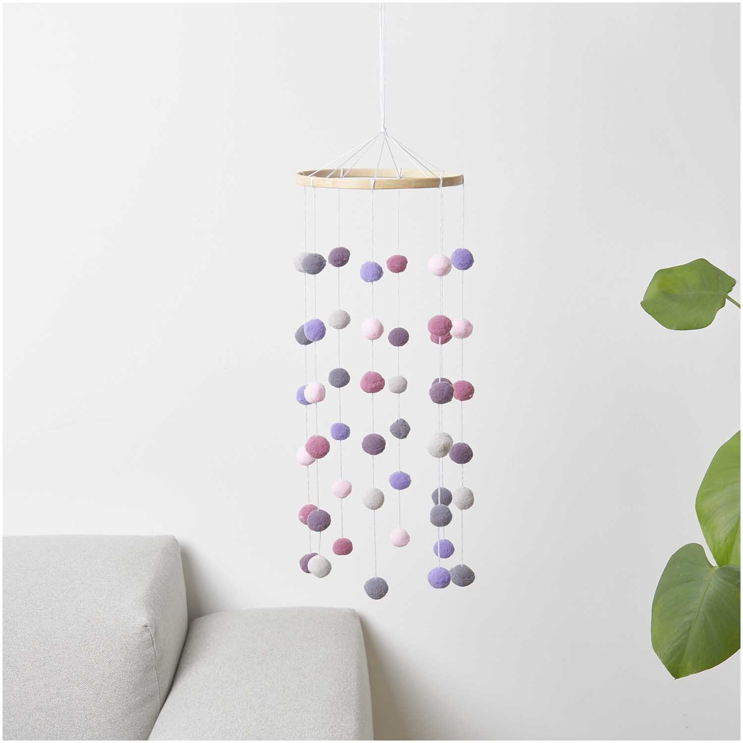 Bastelset Pompon Mobile erdfarben von Rico Design mit bunten Pompons an Holzring, hängend neben Sofa und Pflanze.