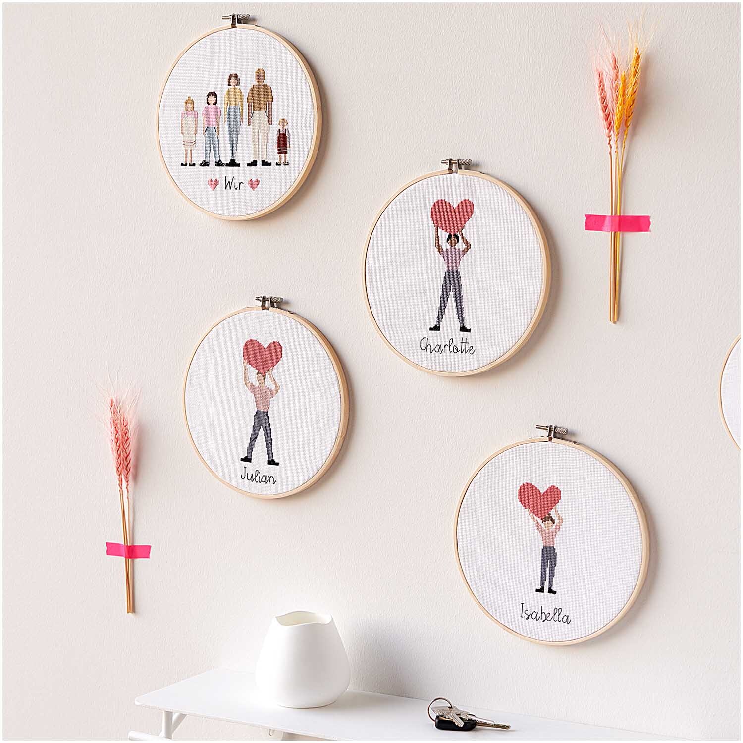 Figurico Stickpackung Family 20cm: Vier Stickrahmen mit Figuren und Herzen an einer Wand, dekoriert mit Trockenblumen.
