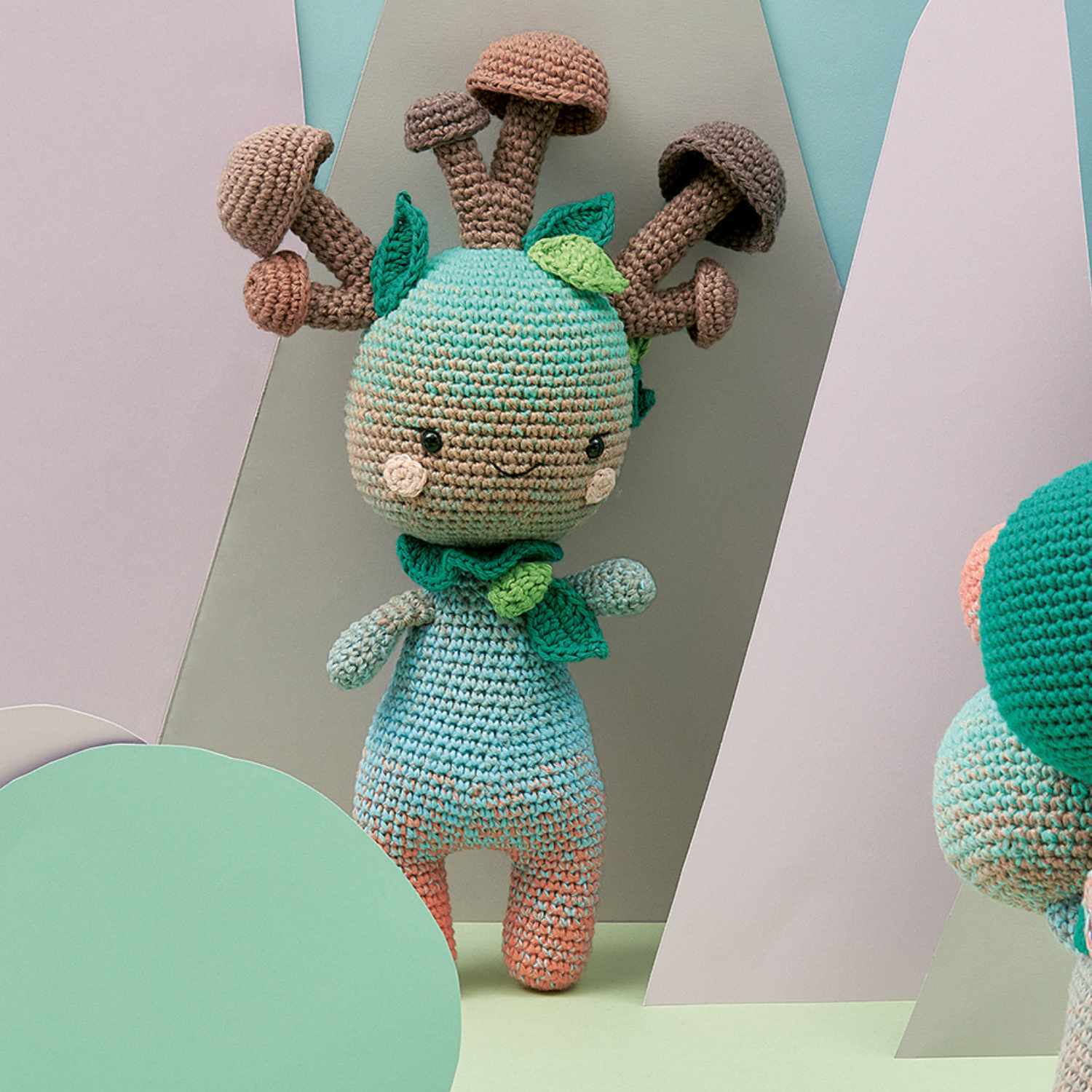 Häkelset Mother-Earth Head: Bunte gehäkelte Figur mit Pilzen und Blättern auf dem Kopf, vor pastellfarbenem Hintergrund.