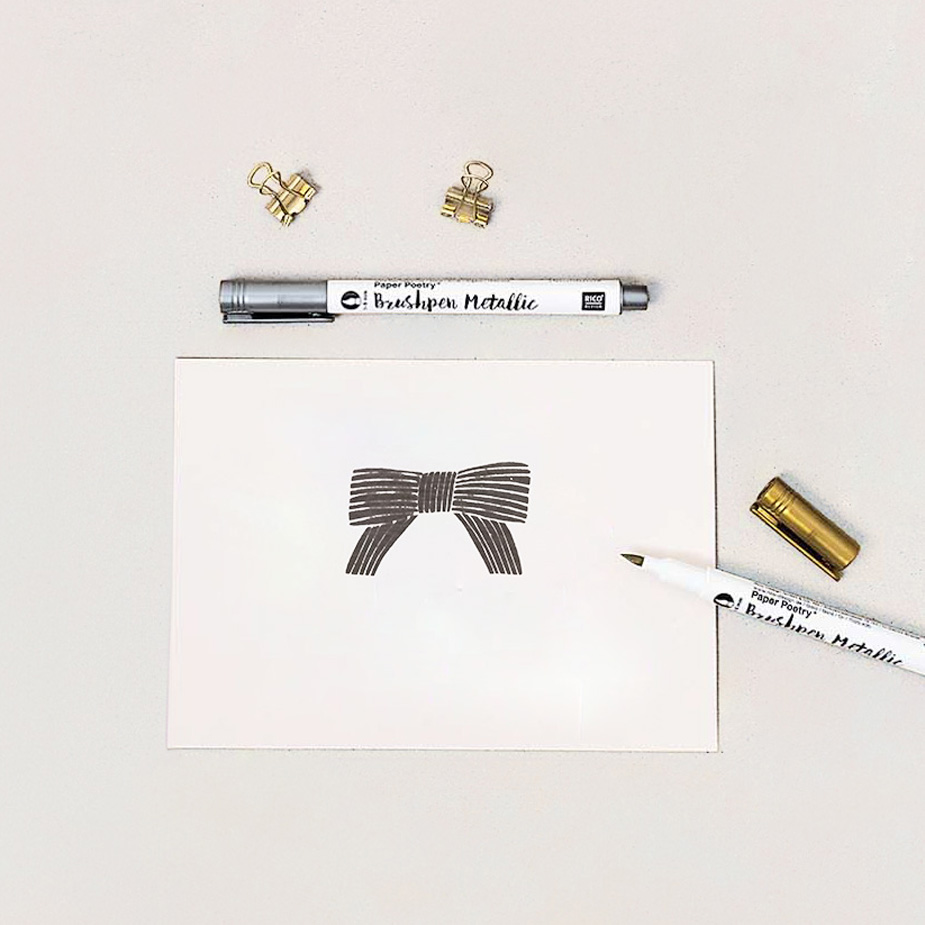 Eine Karte ist mit einer Schleife gestempelt, drumherum liegen zwei metallic Brushpen und zwei goldenen Foldback Klammern. Flatlay von Produkten.