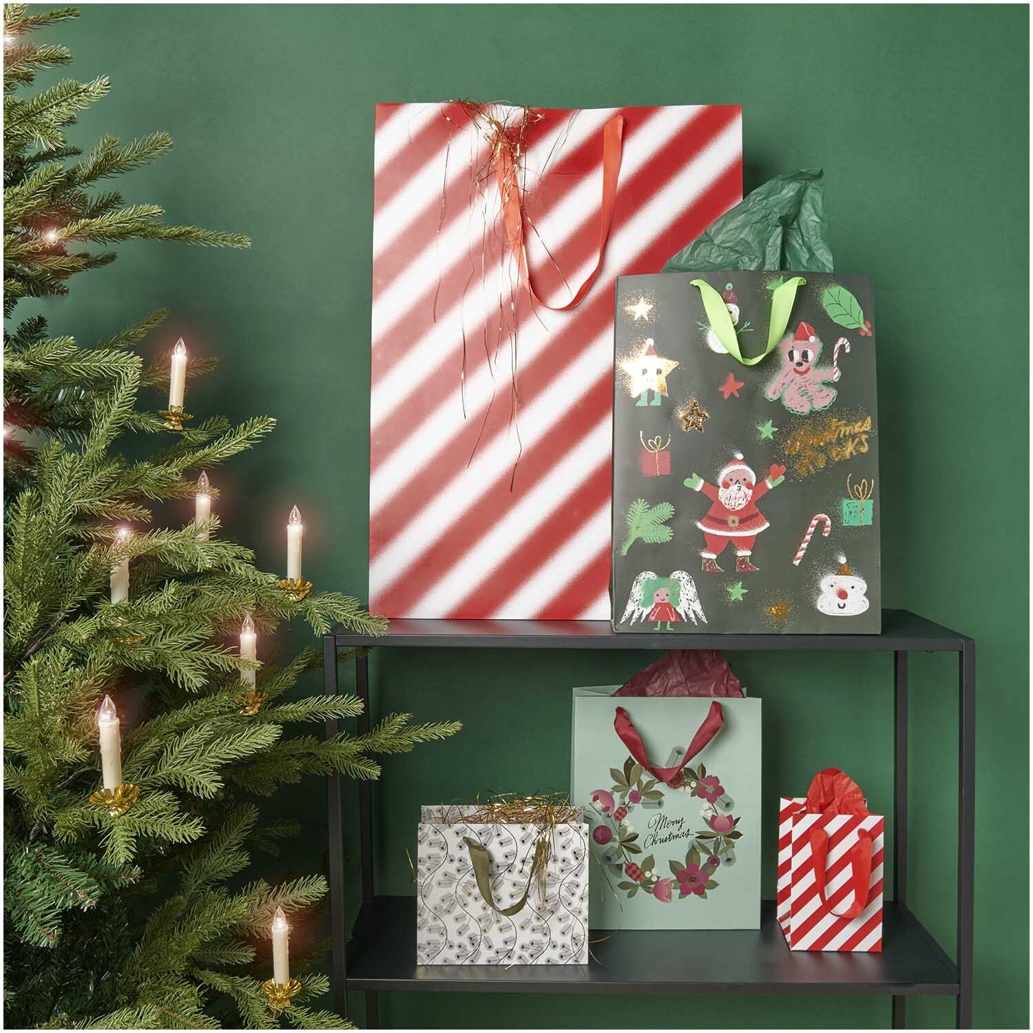 Paper Poetry Geschenktüte Christmas Rocks! in Schwarz mit festlichen Motiven neben einem geschmückten Weihnachtsbaum auf einem Regal.