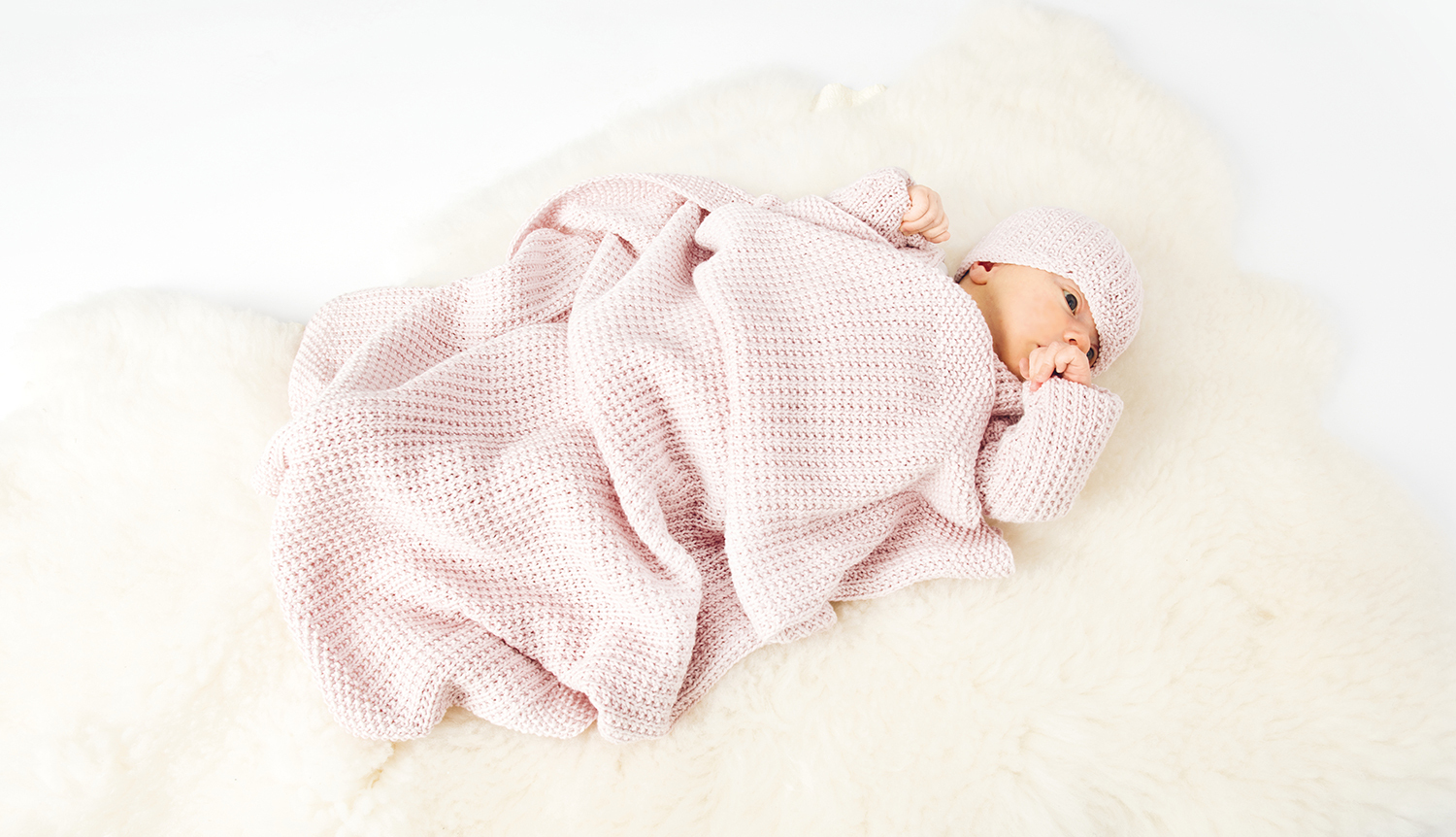 Strickanleitung Baby-Decke: Ein Baby liegt in einer weichen, rosa gestrickten Decke auf einem weißen Hintergrund.
