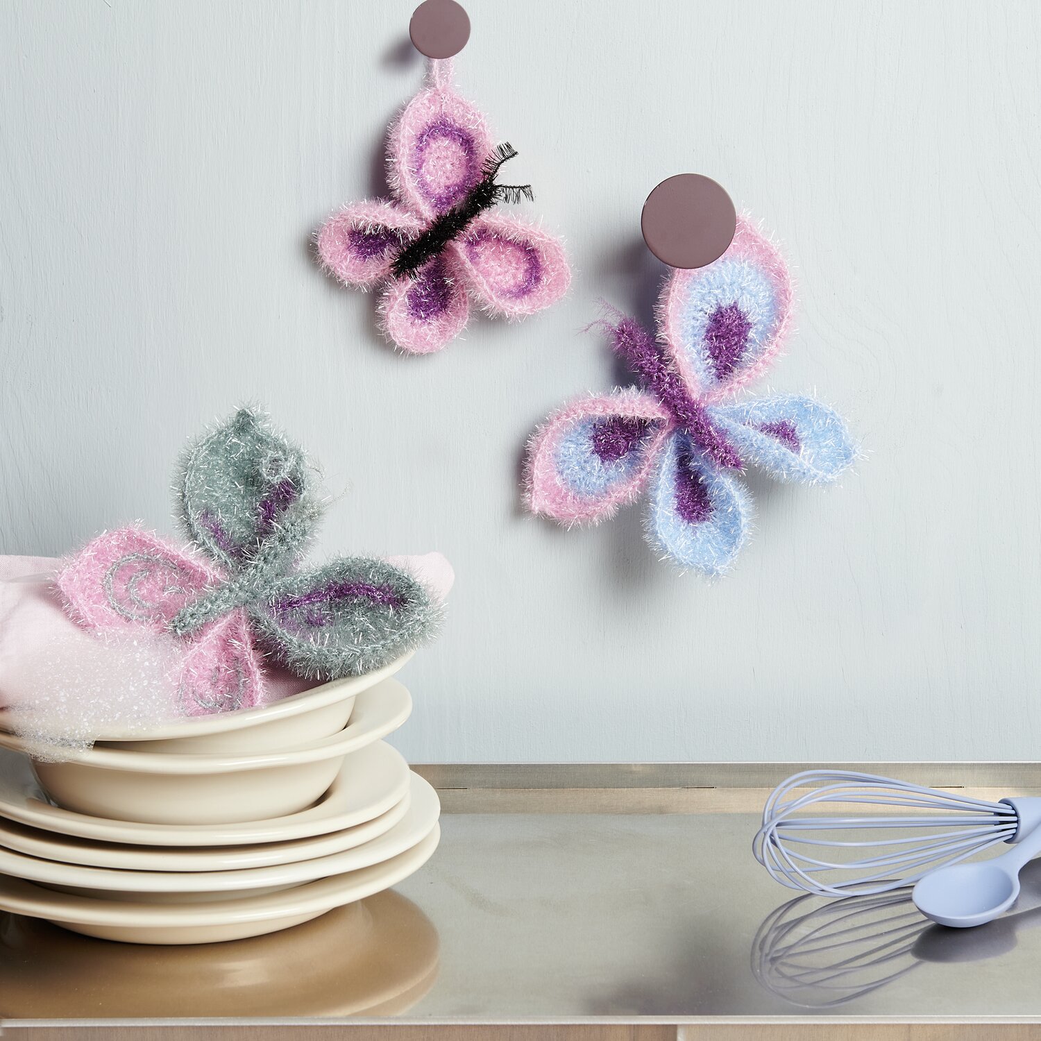 Häkelset Creative Bubble Schmetterling von Rico Design: Zwei gehäkelte Schmetterlinge in Pastellfarben, dekorativ an der Wand und auf Geschirr platziert.