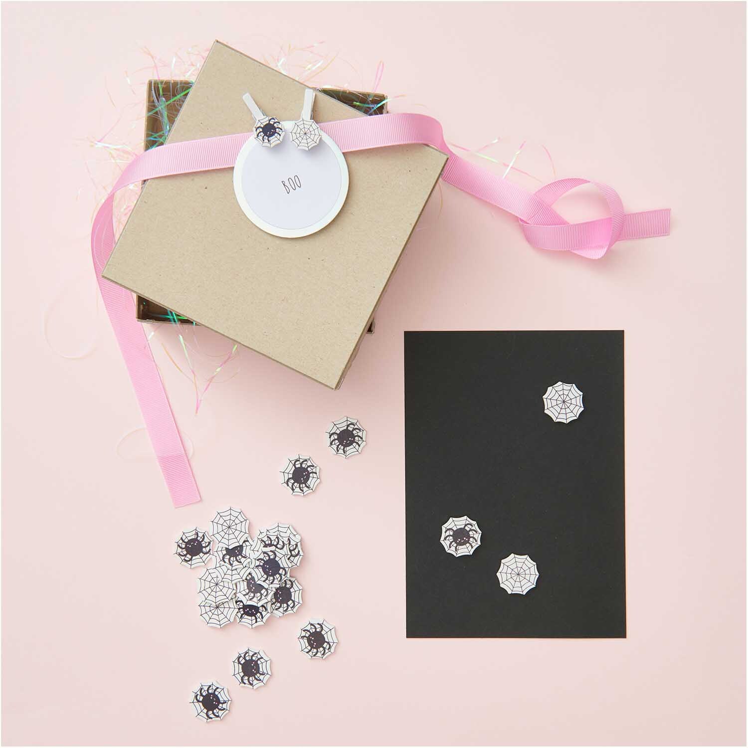 Holzclips Spinnennetz Mix schwarz-weiß 8 Stück auf schwarzem Karton, dekoriert mit rosa Geschenkband. Ideal für kreative Bastelprojekte.