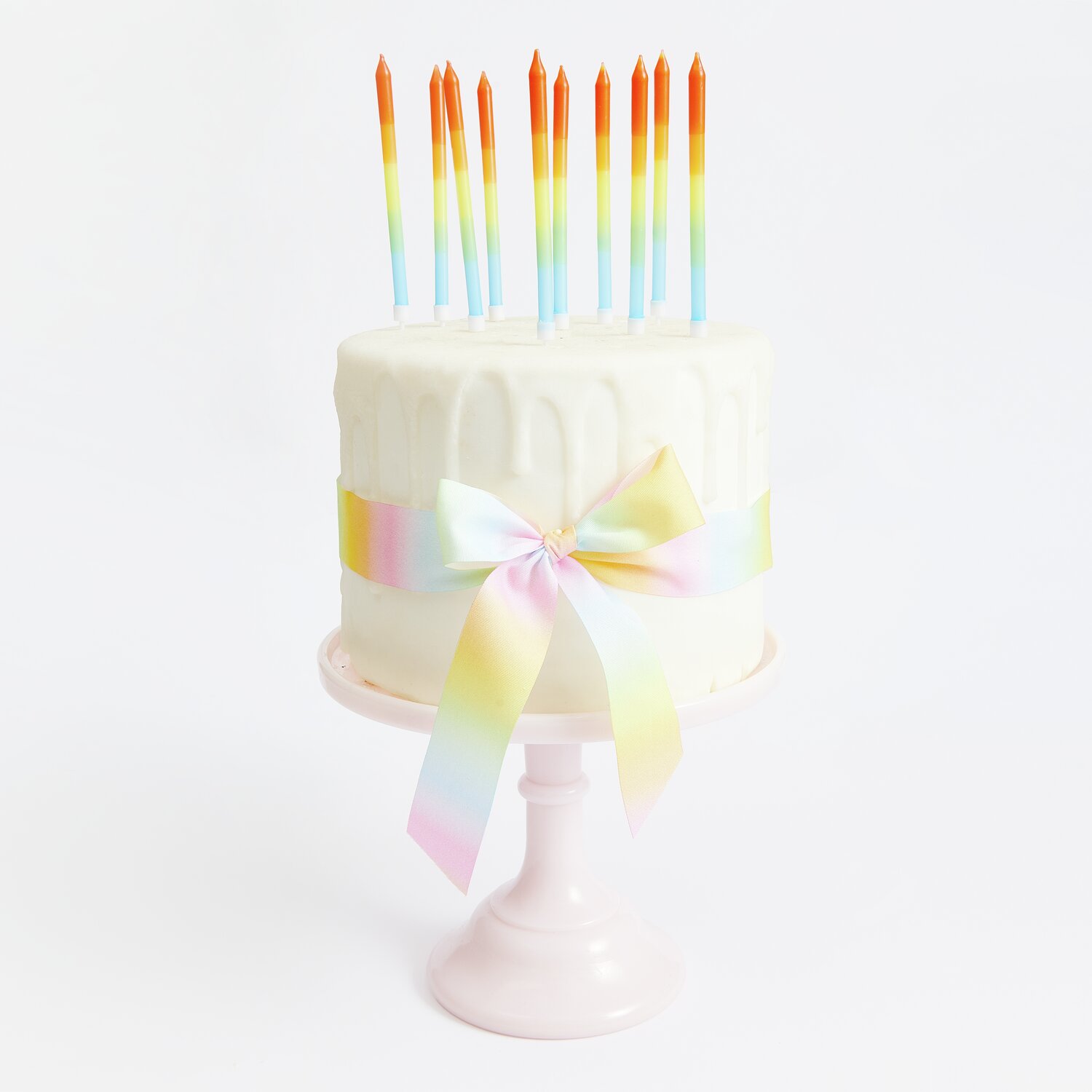 Kerzen Dip Dye Regenbogen auf weißer Torte mit pastellfarbenem Band, präsentiert auf rosa Tortenständer.