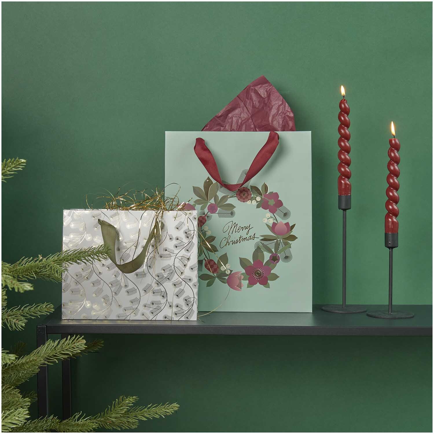 Paper Poetry Geschenktüte mit Tannenzweigen-Muster, dekoriert neben einer weiteren Tüte und roten Kerzen.