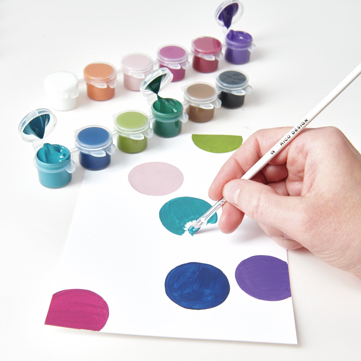 Acrylini Pods: Eine Hand malt mit einem Pinsel auf Papier, umgeben von bunten Farbtöpfen.