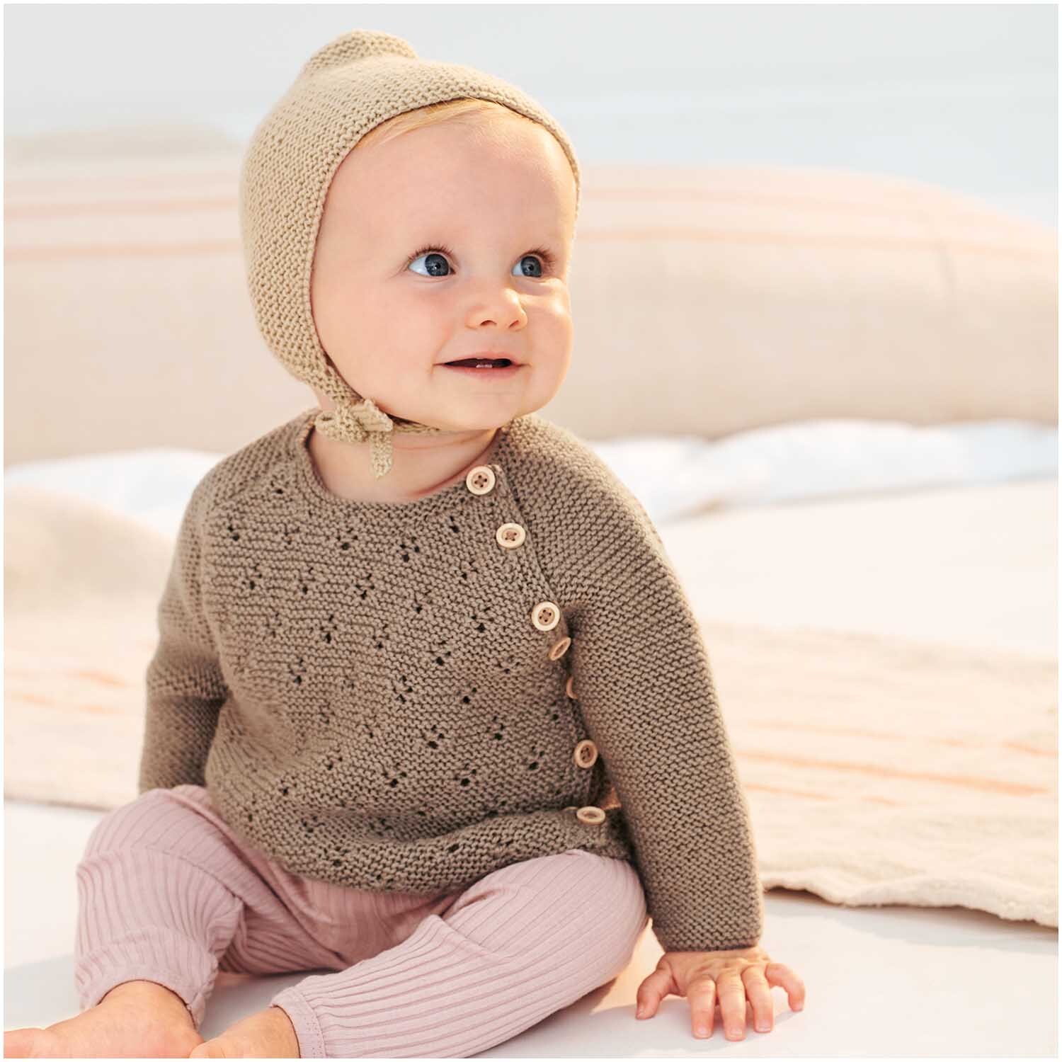 Baby Merino: Ein lächelndes Baby in beiger Strickkleidung von Rico Design, sitzt auf einer Decke.
