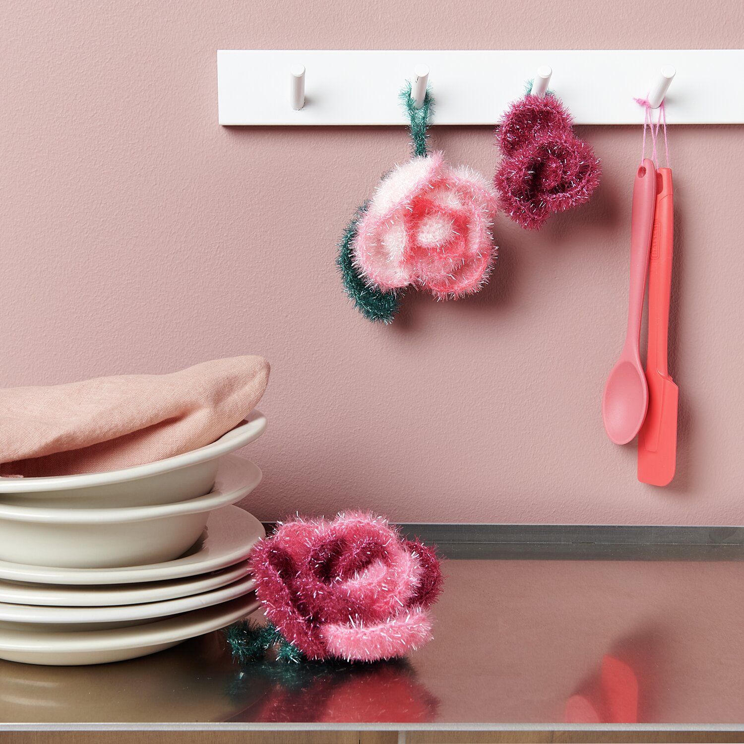 Häkelset Creative Bubble Rose Classic von Rico Design: Rosa und rote gehäkelte Rosen an Haken, mit Geschirr und Küchenutensilien auf Metalloberfläche.