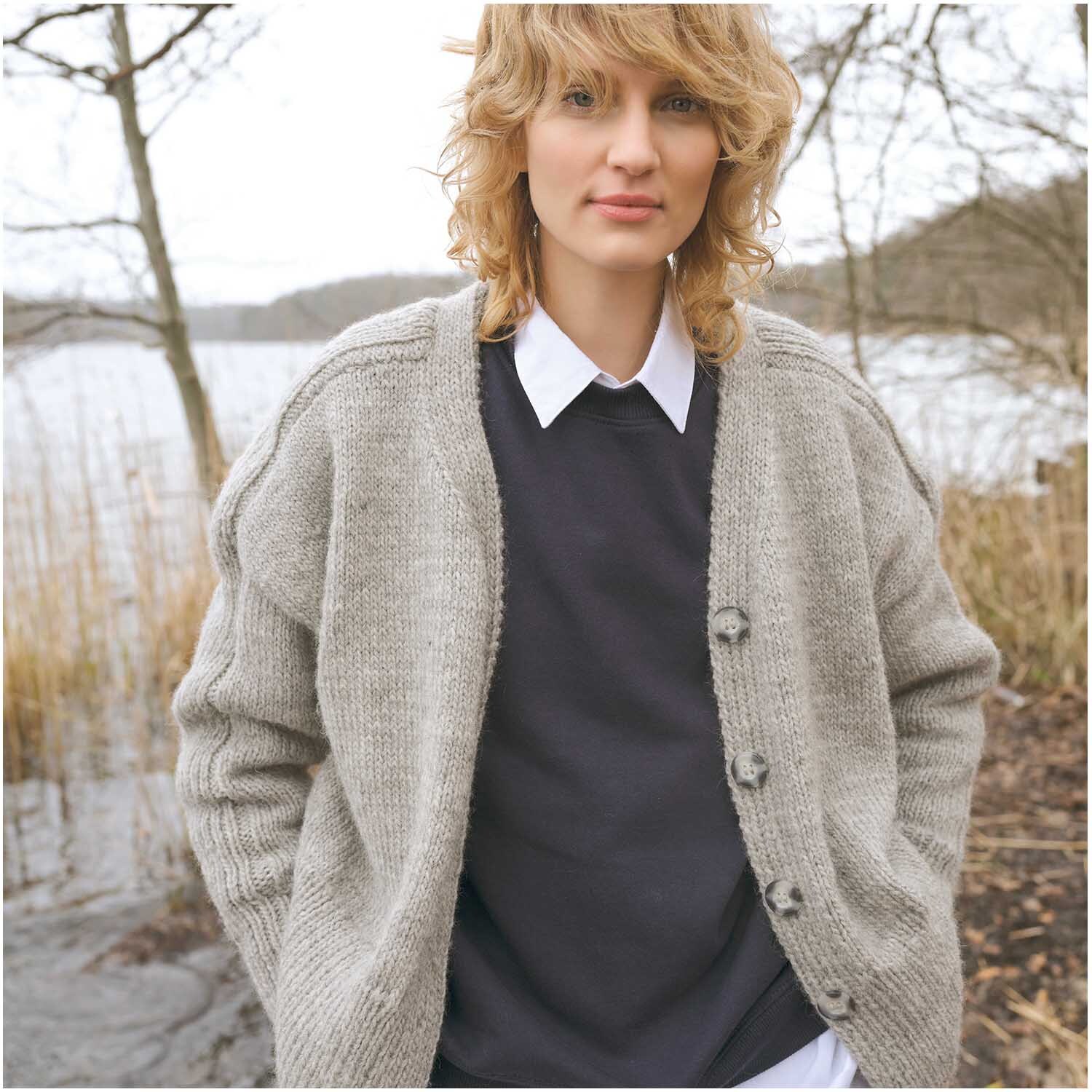 Strickset Jacke Modell 08: Frau in grauer Strickjacke vor einem See mit kahlen Bäumen im Hintergrund.