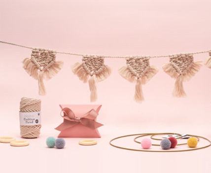 Hängende Makramee-Girlande mit Garn, eine Geschenkschachtel, verschiedene Dekoringe und Verpackung mit Garn-Pompons. Fotosetting rosa Wand.