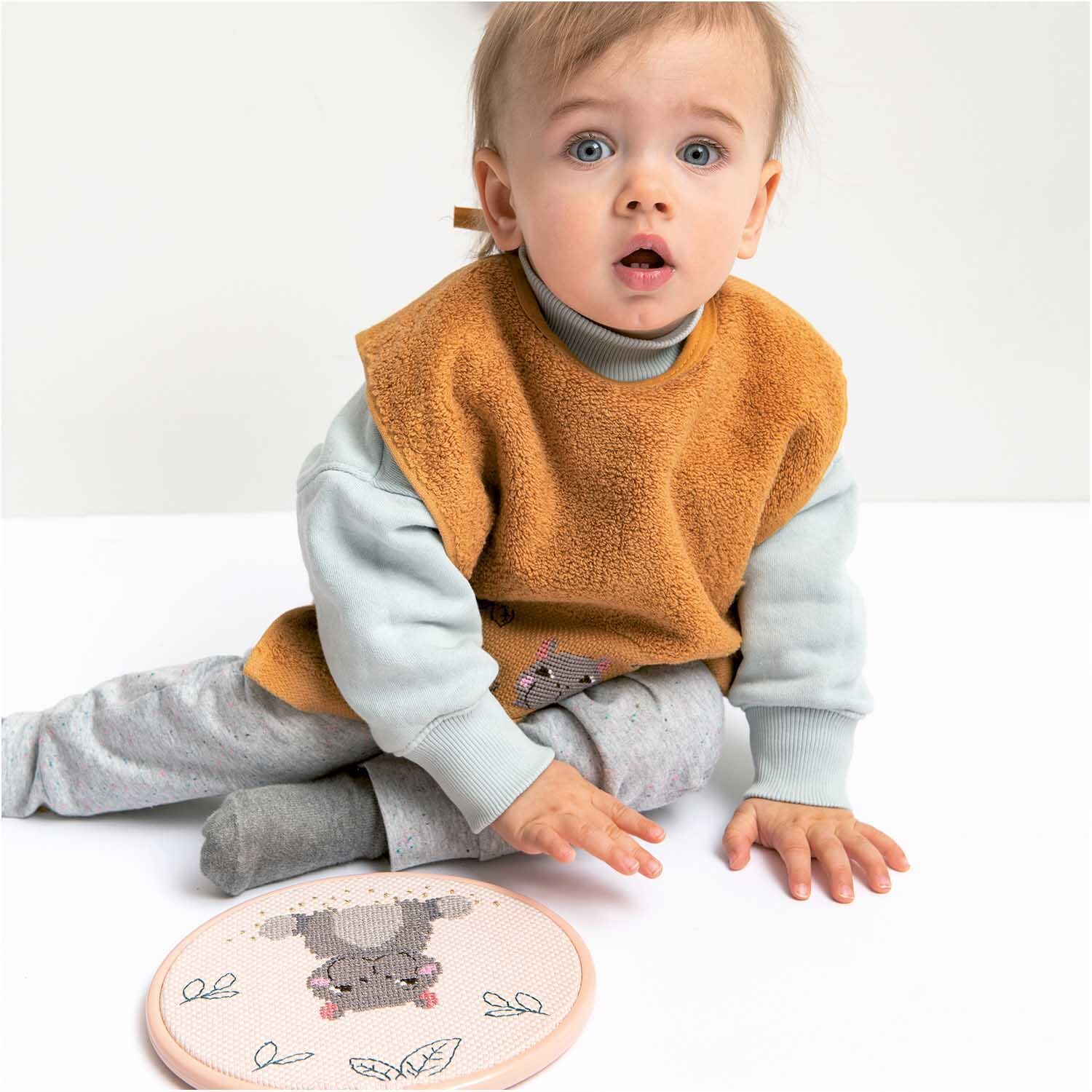 Stickring aus Kunststoff puder 17,8cm: Ein Baby sitzt auf dem Boden, trägt einen orangefarbenen Latz und spielt mit einem rosa Stickring von Rico Design.
