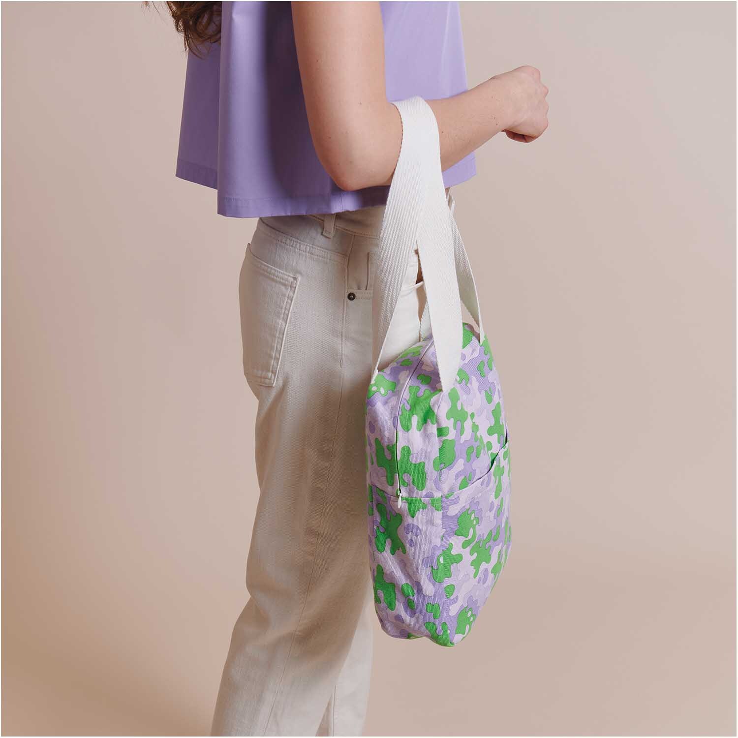 Stoffabschnitt Canvas Camouflage grün-lila 50x140cm: Frau trägt Tasche aus mehrfarbigem Camouflage-Stoff von Rico Design.
