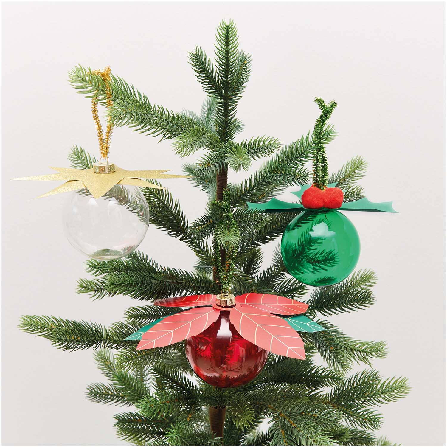 Paper Poetry Christbaumkugel-Topper Stern in Gold von Rico Design auf einem Tannenbaum. Drei Kugeln in Rot, Grün und Transparent mit dekorativen Elementen.