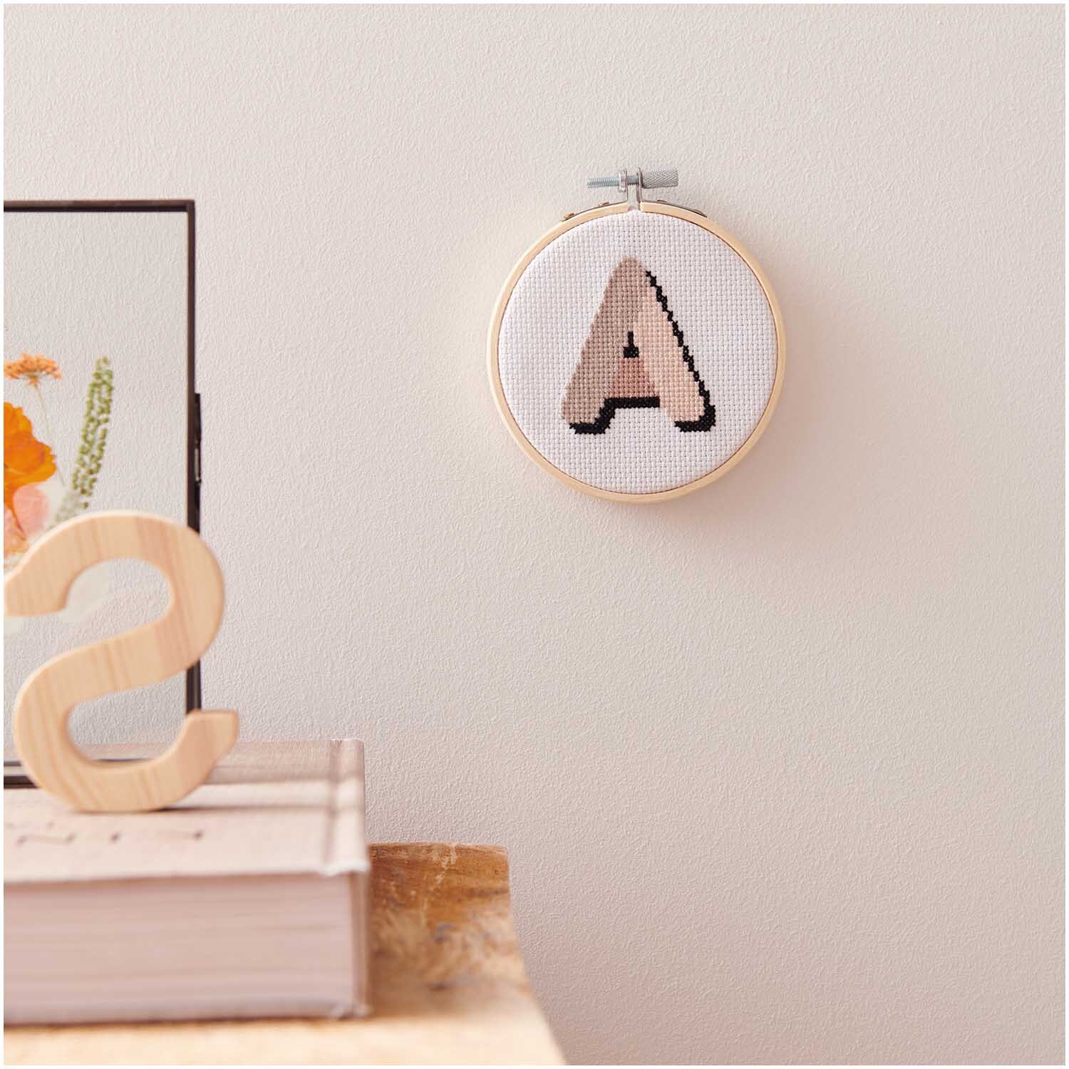Stickpackung Mini Kit Alphabet Bild Ø 10cm zeigt ein gesticktes A in Holzrahmen an einer Wand.