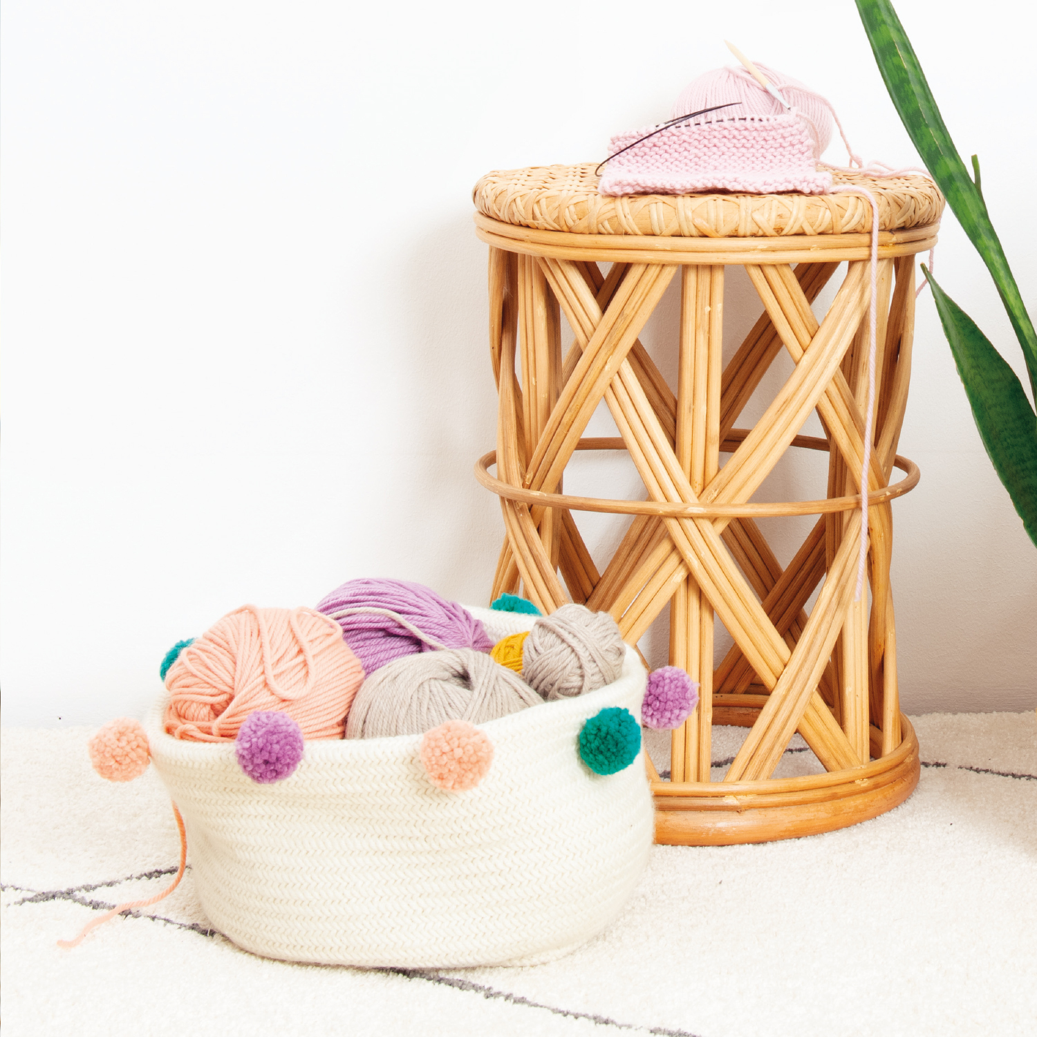 Strickanleitung Korb aus Essentials Mega Wool Chunk: Ein Korb mit bunten Pompons, gefüllt mit Garn, neben einem geflochtenen Hocker.