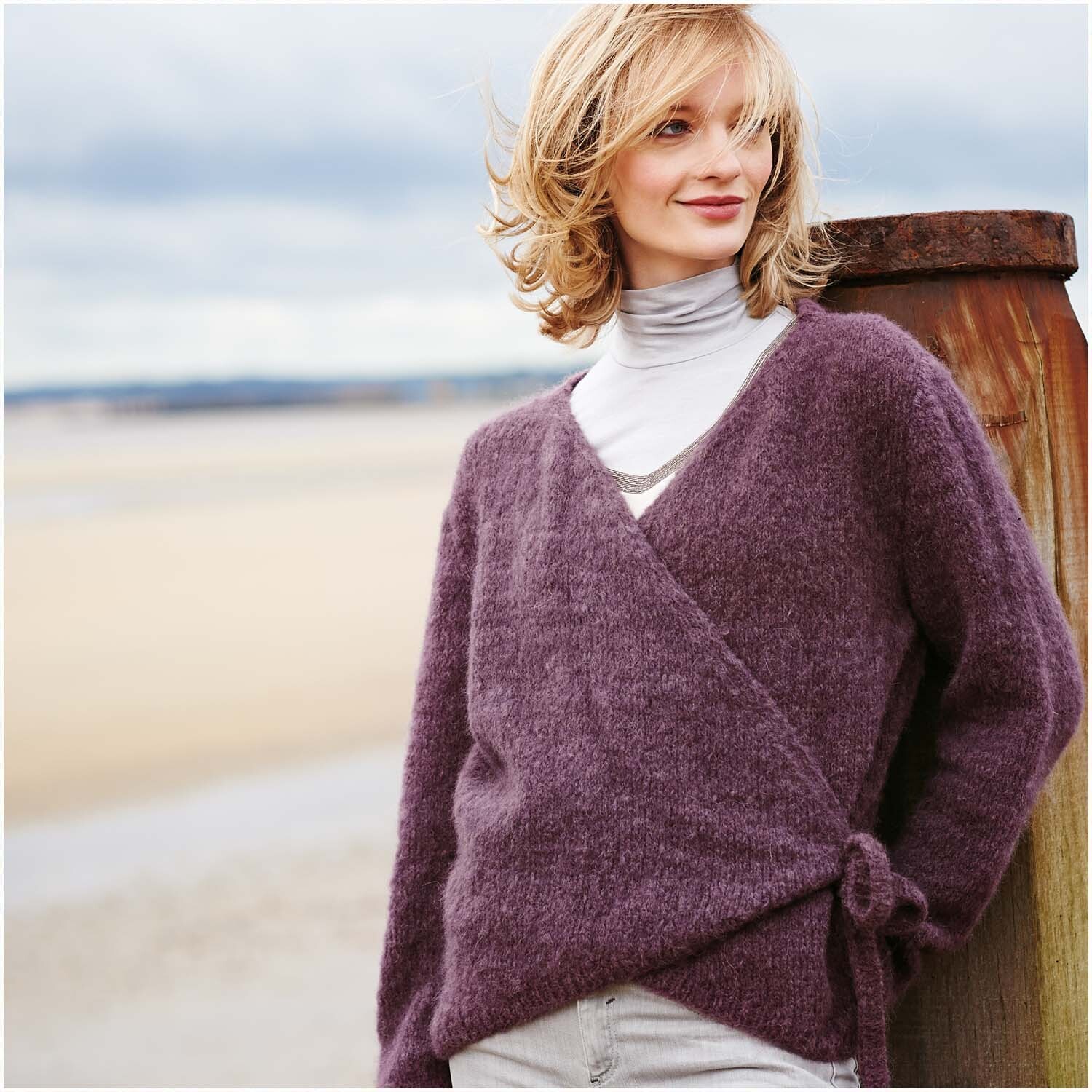 Luxury Volumi: Frau in lila Wickeljacke am Strand, lächelnd, mit grauem Rollkragenpullover.