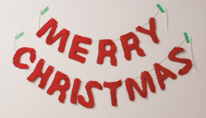 Strickanleitung Merry Christmas Girlande: Rote gestrickte Buchstaben formen den Schriftzug "Merry Christmas" an einer Wand.