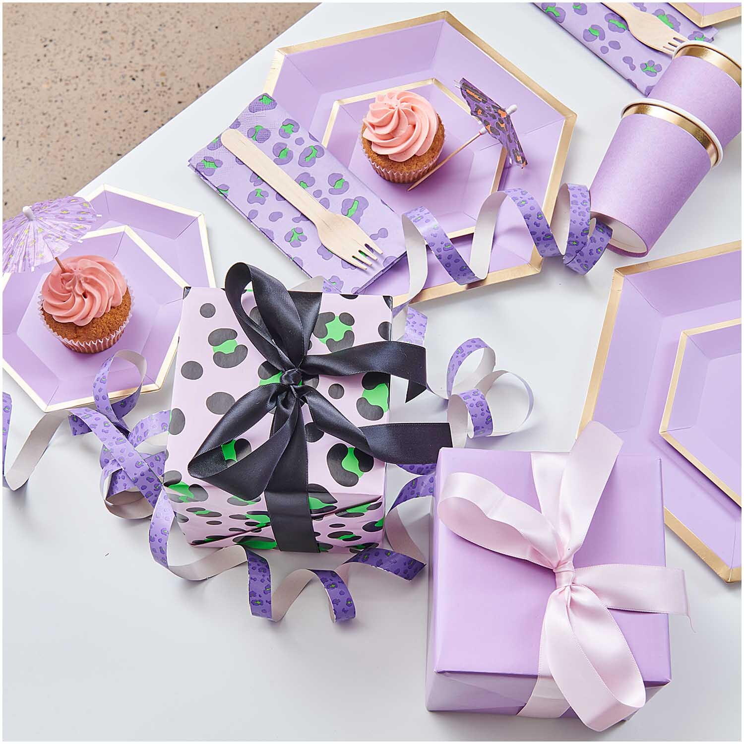 Paper Poetry Geschenkpapier Acid Leo rosa umhüllt Geschenke, umgeben von lila Partygeschirr und Cupcakes.