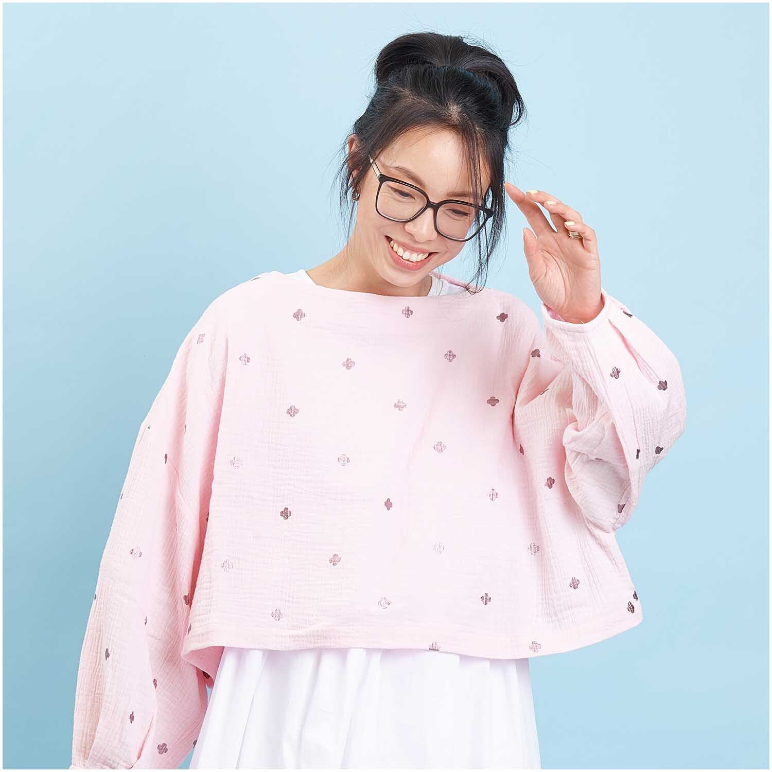Stoffabschnitt Krinkelmusselin Blüten rosa: Frau in rosa Oberteil mit Blütenmuster, lächelnd vor blauem Hintergrund. Marke: Rico Design.