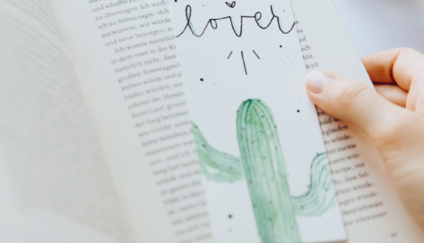 Anleitung Lesezeichen mit Kaktus: Hand hält ein Buch mit einem weißen Lesezeichen, das einen grünen Kaktus zeigt.
