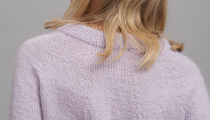 Strickanleitung Pullover aus Essentials Fine Fine Merino: Frau trägt lila gestrickten Pullover, Rückansicht, weiches Material, ideal für kühle Tage.