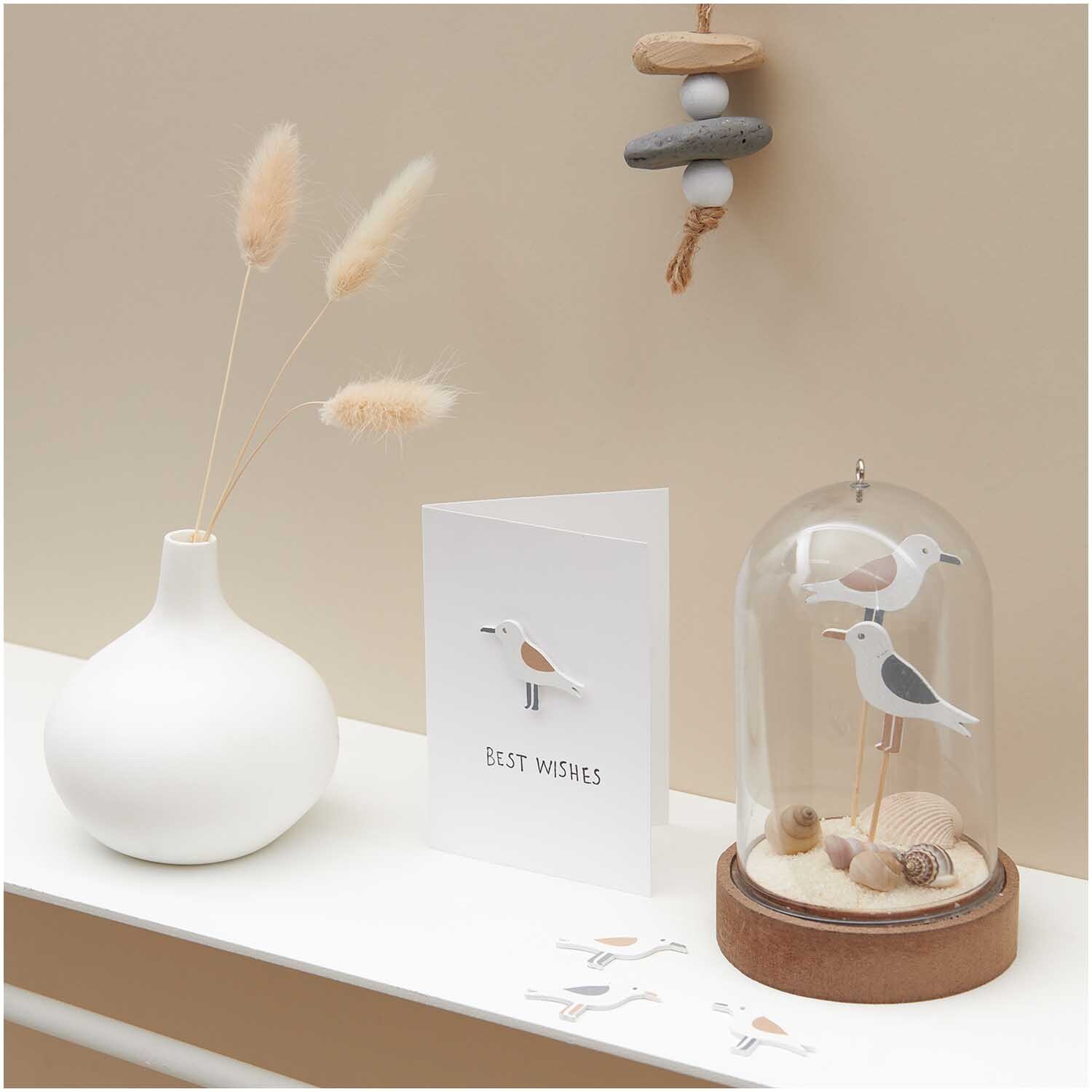 Holzstreu Möwe natur 35x26mm 36 Stück von Rico Design: Dekorative Möwenfiguren unter Glas mit Muscheln, neben einer weißen Vase und einer Grußkarte.