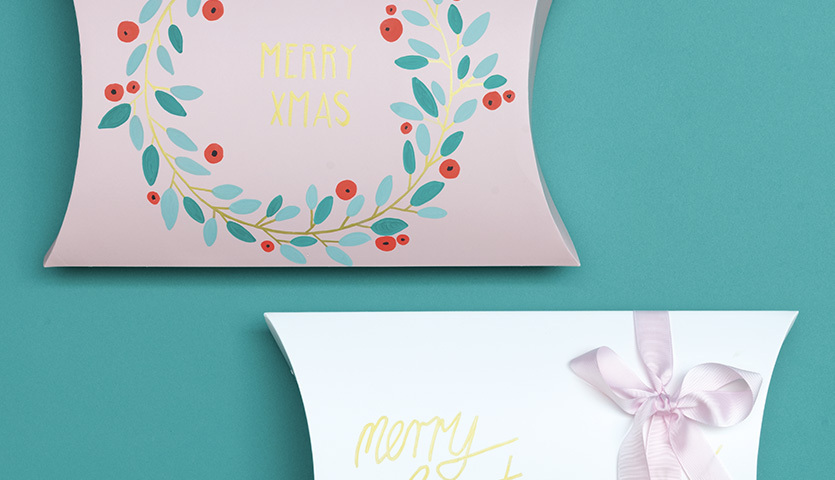 Anleitung Geschenkschachteln mit Kreidemarkern gestalten: Zwei dekorierte Schachteln, eine mit Blumenkranz, die andere mit Schleife, auf türkisfarbenem Hintergrund.