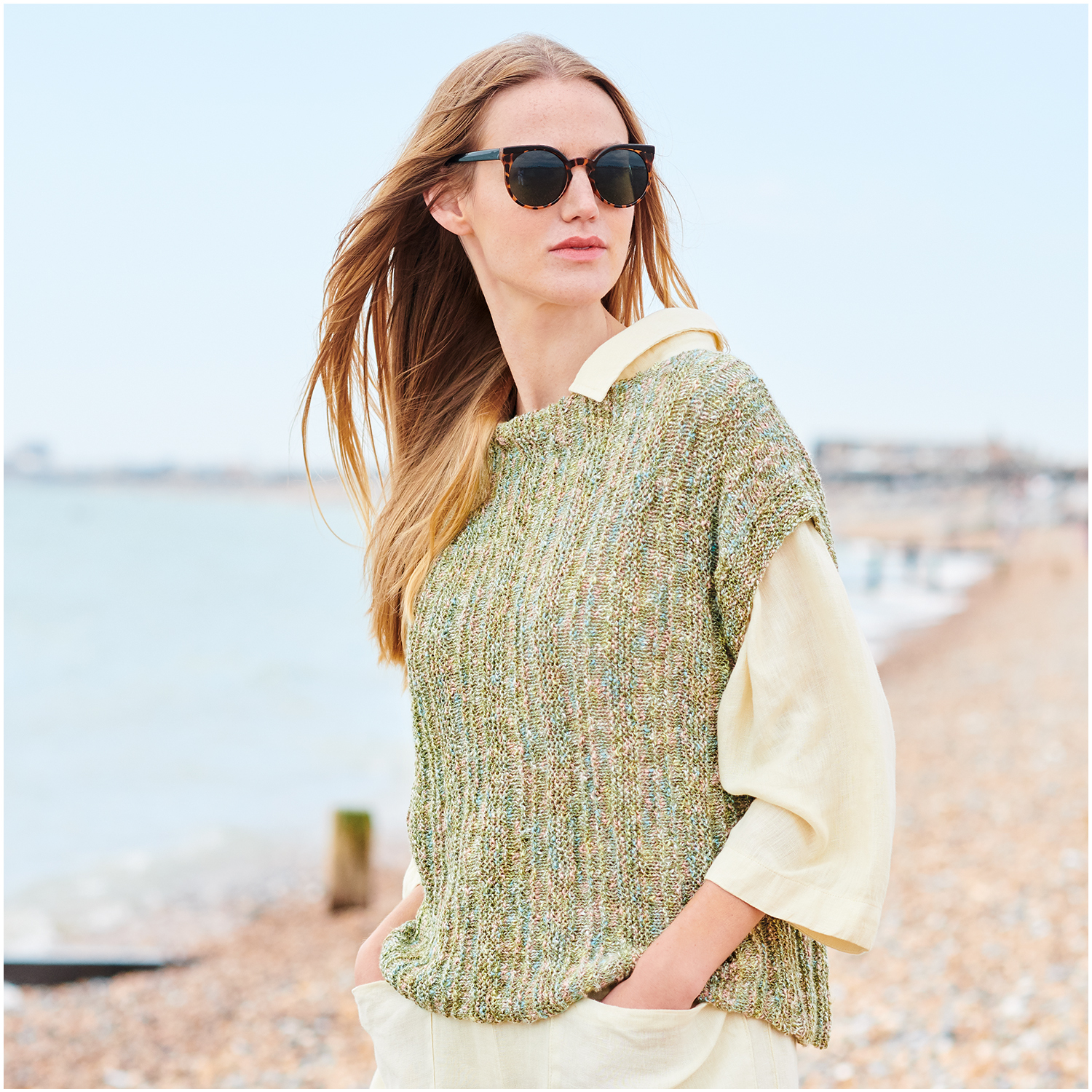 Strickset Top Modell 26 aus Lovewool Nr. 18: Frau am Strand in grünem Stricktop und beiger Bluse, trägt Sonnenbrille.