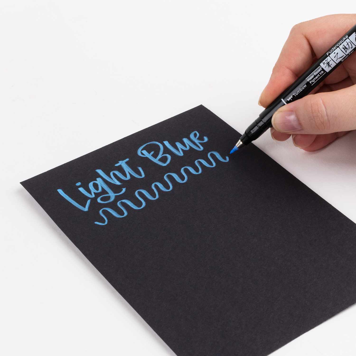 Fudenosuke Brush Pen Pastel schreibt in hellblauer Farbe auf schwarzem Papier. Hand hält Tombow-Stift.