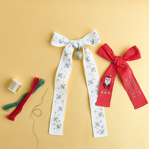 Eine weiße Schleife mit Sternen und Tannenzweigen bestickt, daneben eine kleinere rote Schleife mit Weihnachtsmann und Schriftzug bestickt. Stickset aus der Kollektion Put A Bow On It.