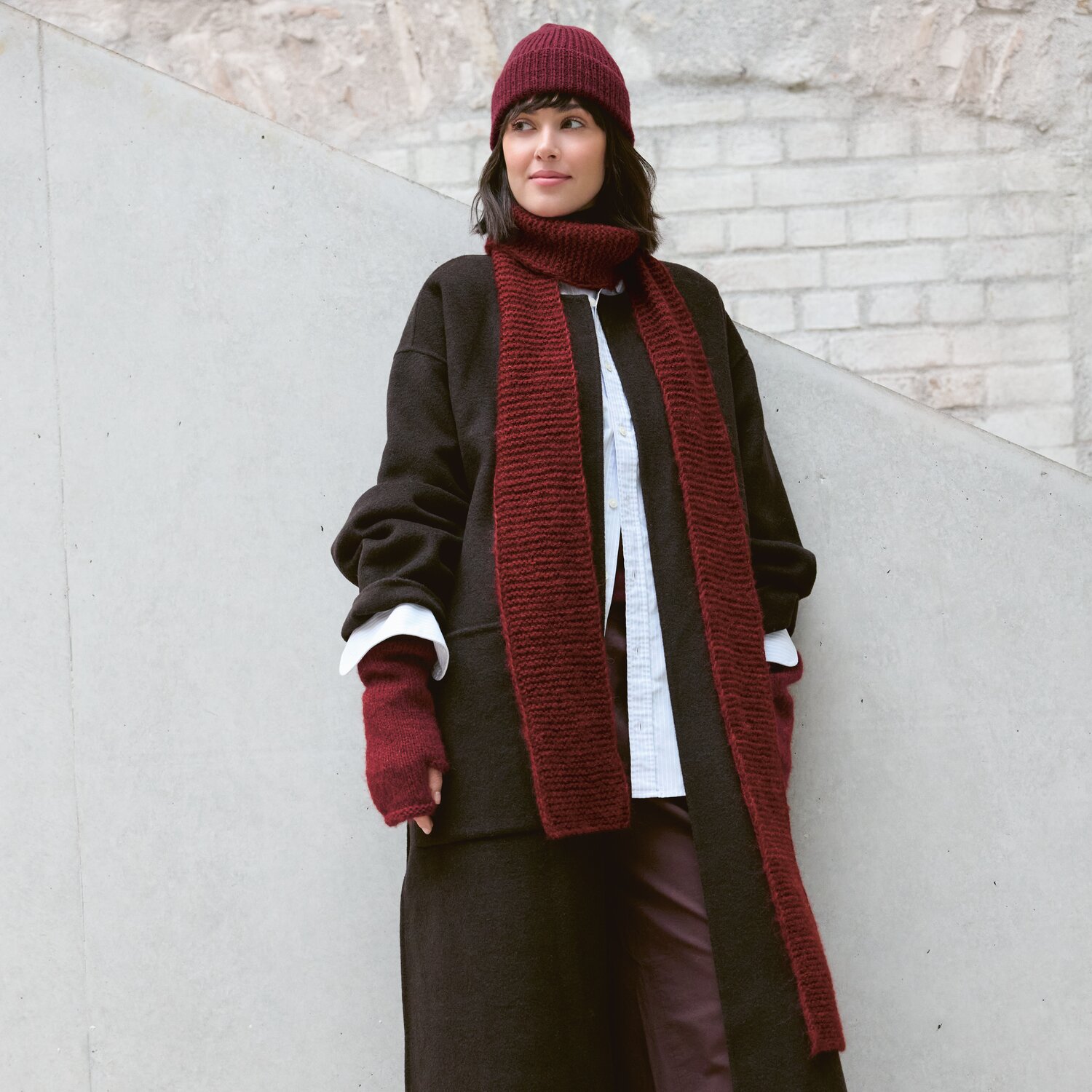 Strickset Schal Modell 17 aus Luxury Knits: Frau trägt roten Schal, Mütze und Handschuhe, kombiniert mit dunklem Mantel vor heller Wand.