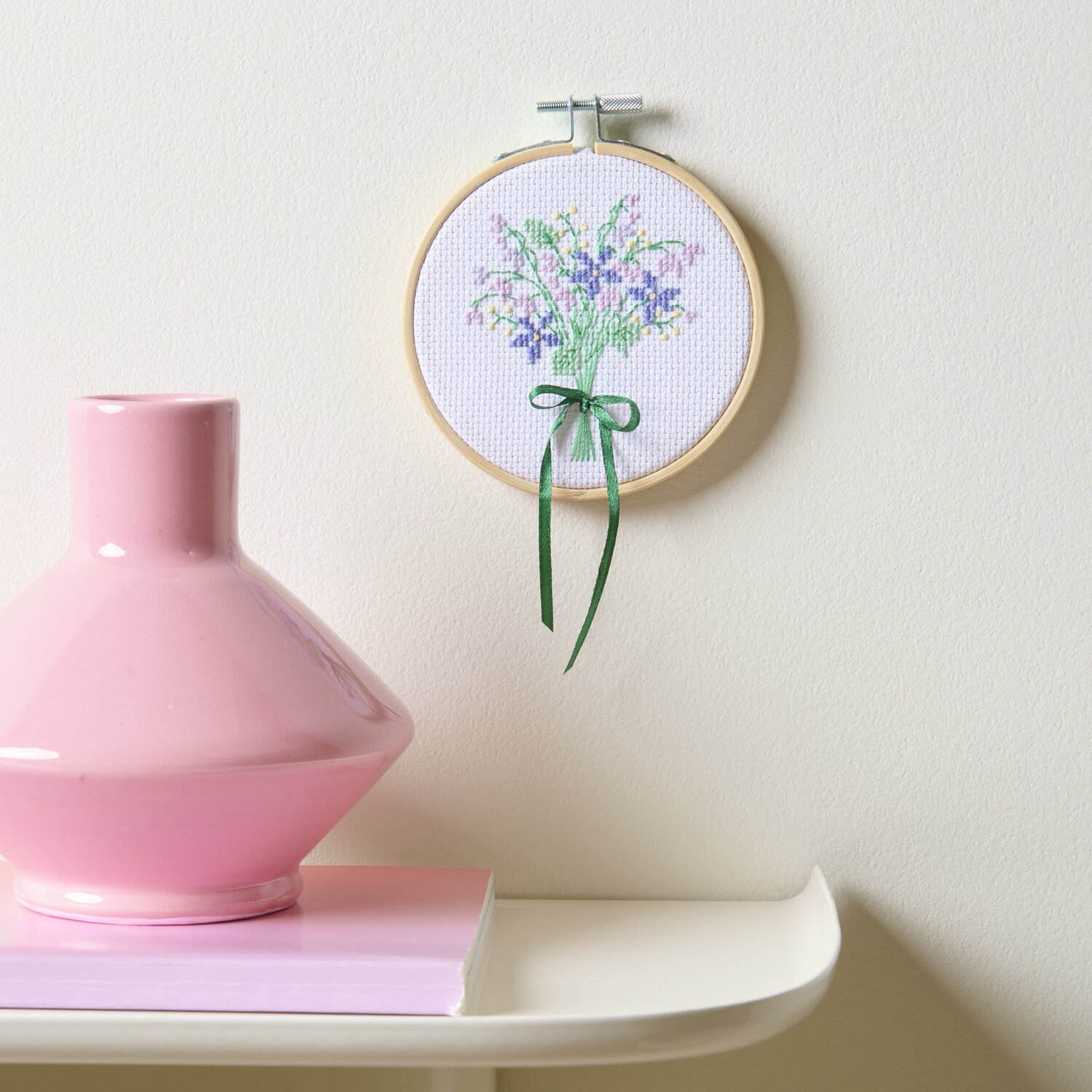 Stickpackung Gezählter Kreuzstich Blumenstrauß: Rundes Stickbild mit Blumenmotiv, Ø10cm, an weißer Wand, neben rosa Vase auf Tisch.